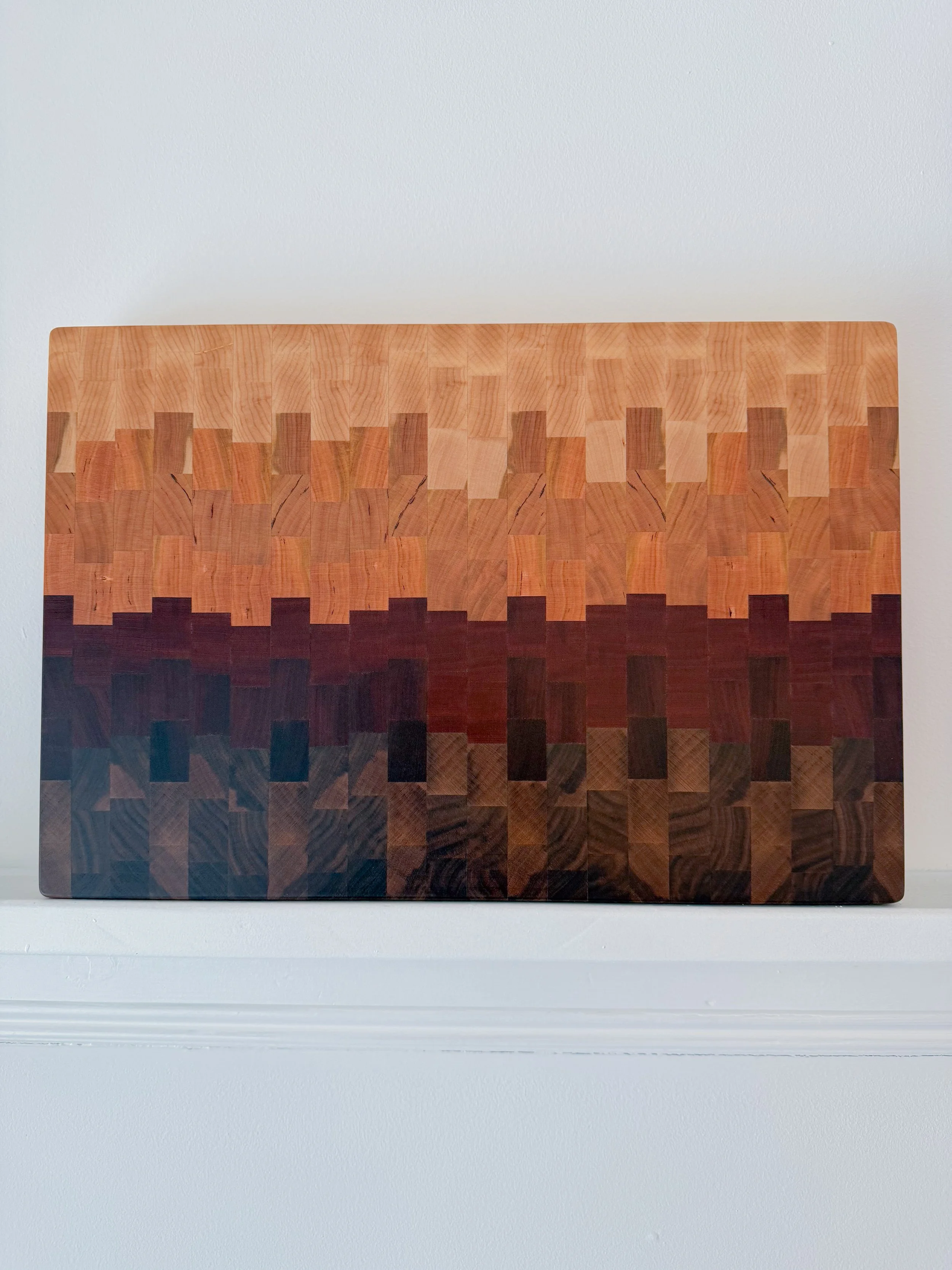 A wooden art piece with a pixelated sunset pattern, featuring gradations from light wood tones at the top to dark brown at the bottom, mounted on a white wall.