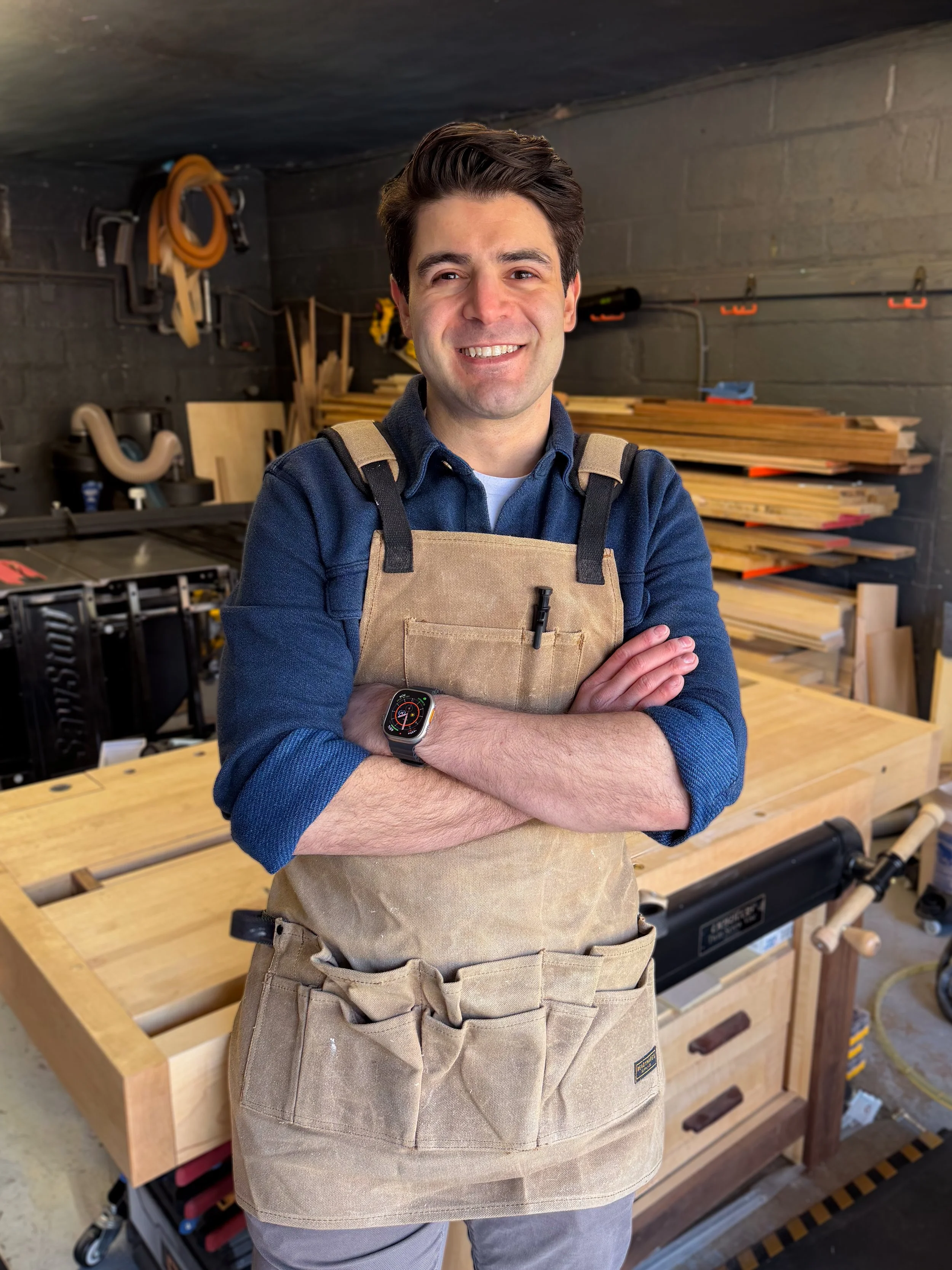 A man in a workshop wearing a beige apron, standing with arms crossed, smiling, with woodworking tools and materials in the background.