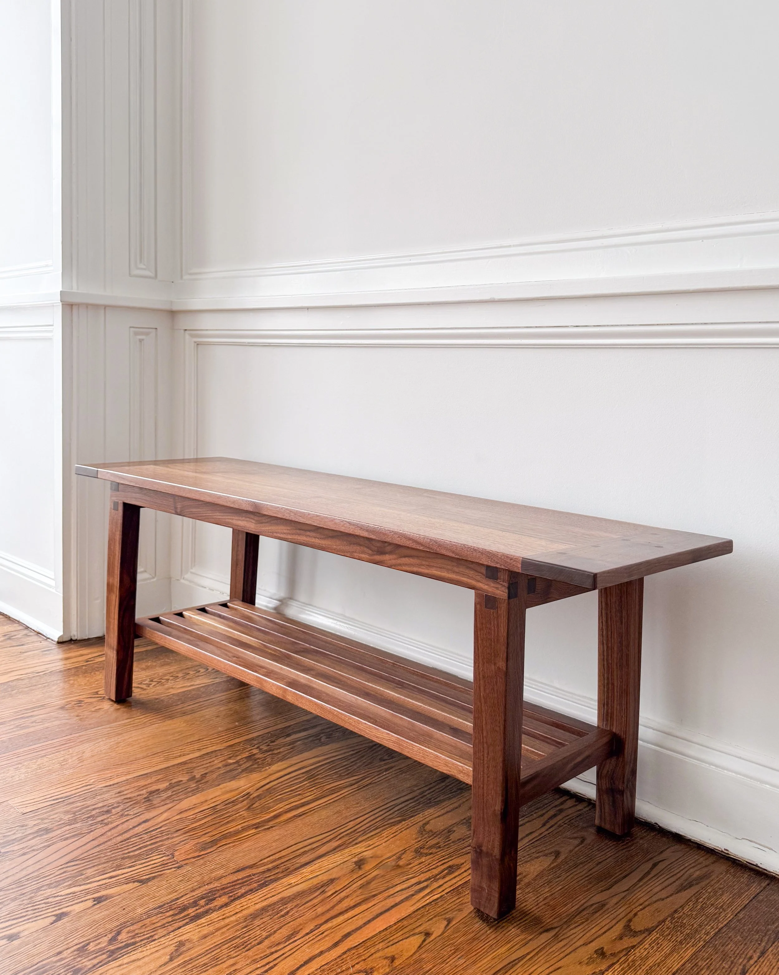Wooden bench with slatted lower shelf against white wall with decorative paneling, hardwood floor.