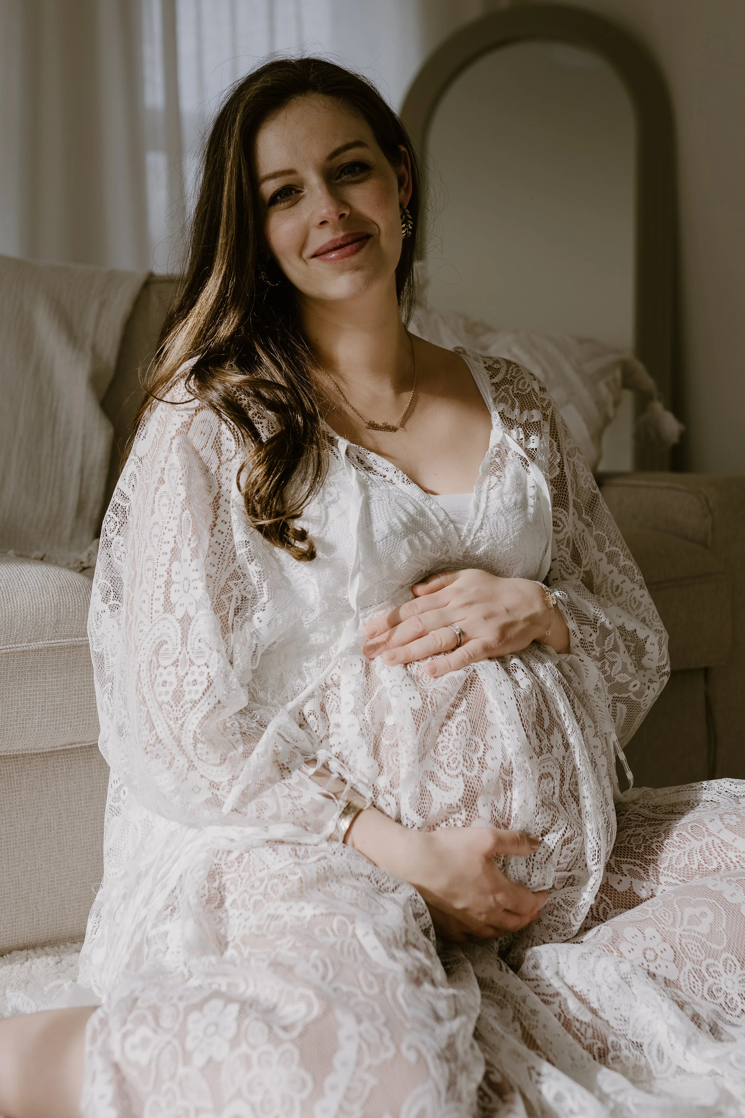 A pregnant woman with long brown hair, smiling softly, sitting on a bed in a sunlit room, wearing a white lace dress that reveals her baby bump in Surrey.