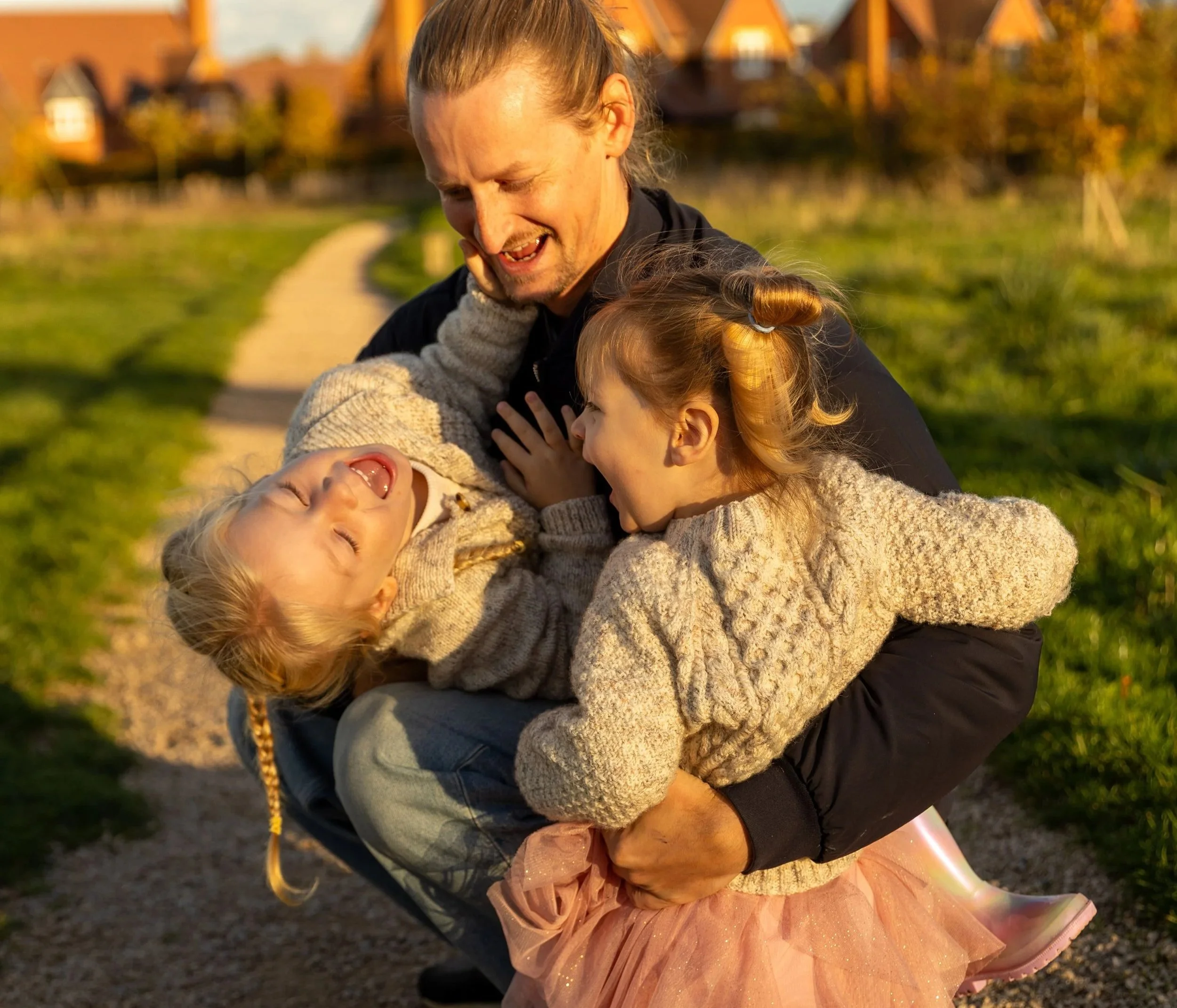A man hugging two young girls outside on a sunny day, all laughing and enjoying a playful moment on a park path.