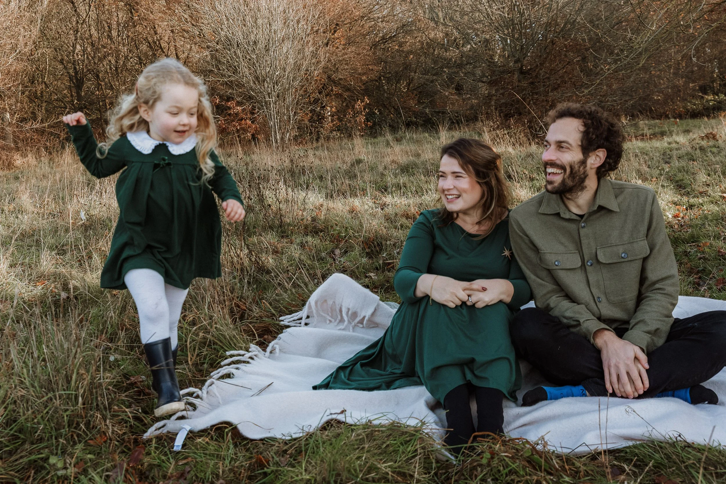 A young girl in a green dress and white tights playing on a picnic blanket with a woman and a man, both smiling, sitting on the blanket outdoors in a grassy field with trees in the background.