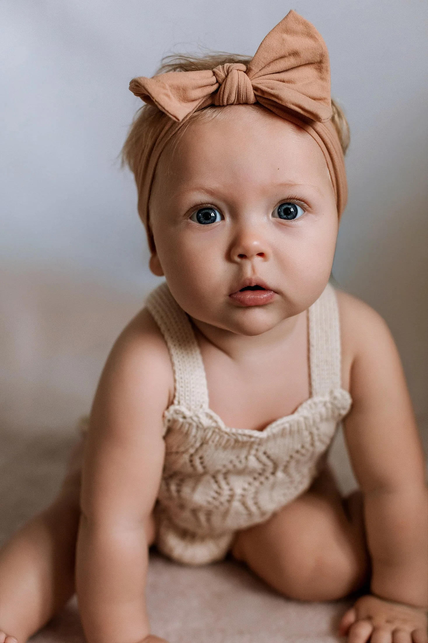 A baby girl with blue eyes, wearing a beige knitted outfit and a tan headband with a bow, sitting on a soft surface and looking directly at the camera.