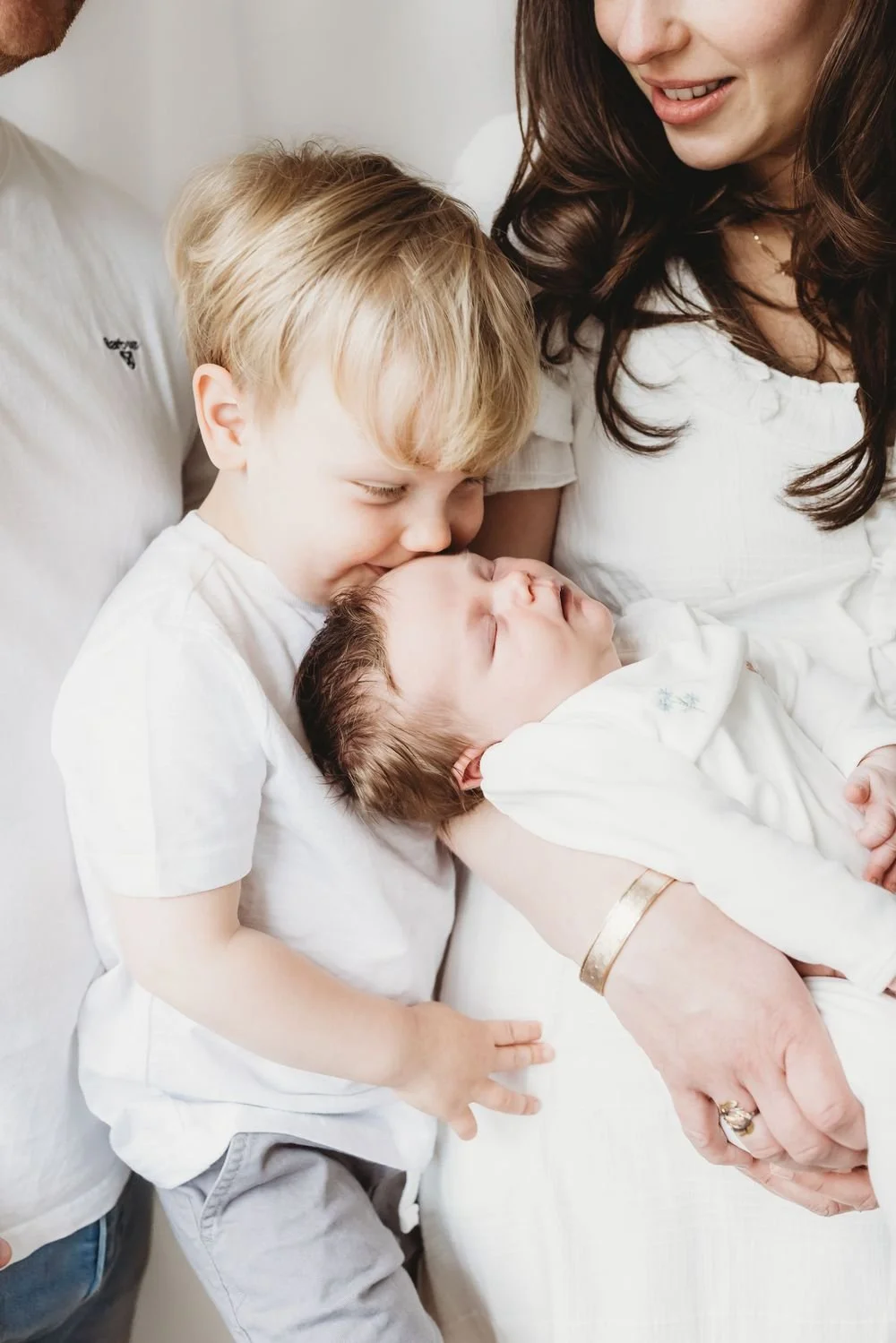 A young boy kissing a sleeping baby who is lying in a woman's arms, with another person partially visible in the background in Surbiton.