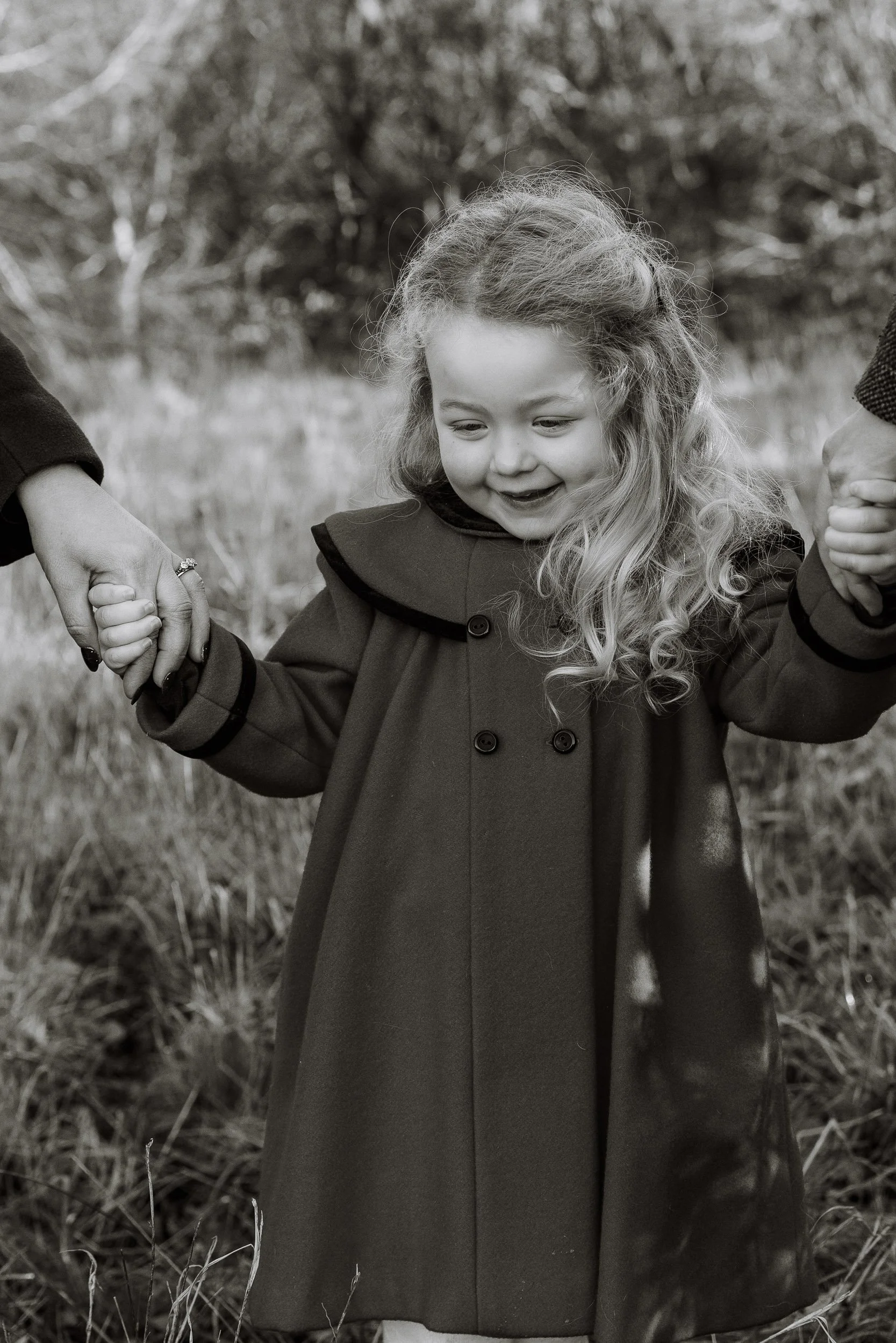 A young girl with curly hair smiling while holding hands with two adults outside in a grassy area.