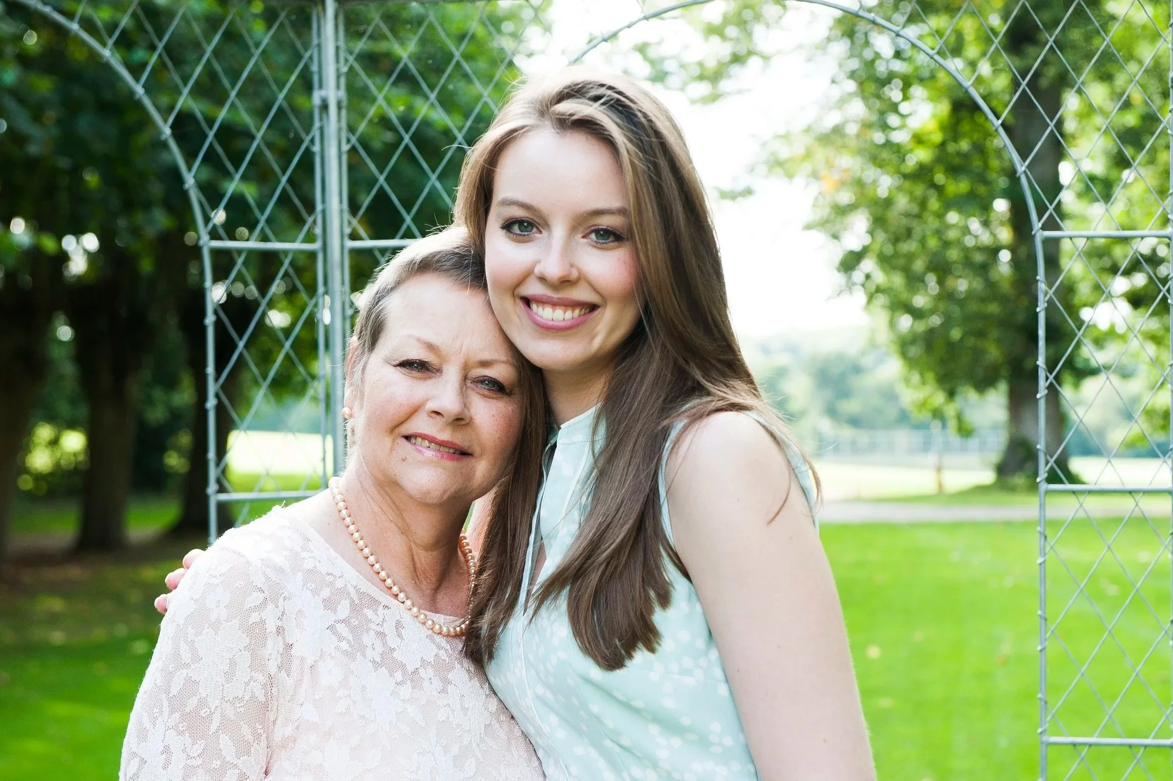 Two women, an older woman and a younger woman, standing close together outdoors, smiling for the camera. They are in a park with green trees and grass, and a metal fence in the background in Berkshire.