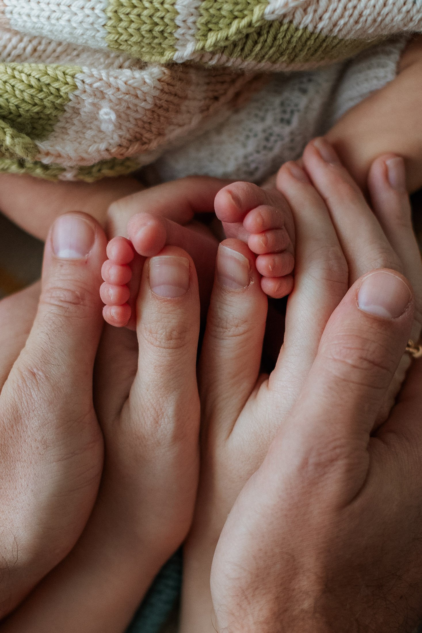 Close-up of adult hands gently holding the tiny feet of a newborn baby, with the baby wearing a knit sweater or blanket.