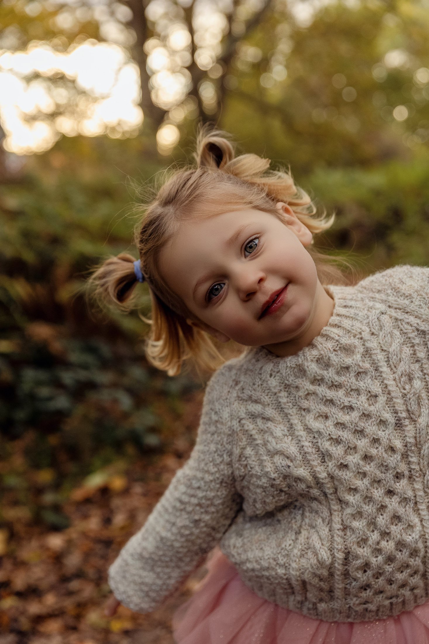 Young girl with pigtails wearing a beige knitted sweater, outdoors in fall forest, smiling at camera.