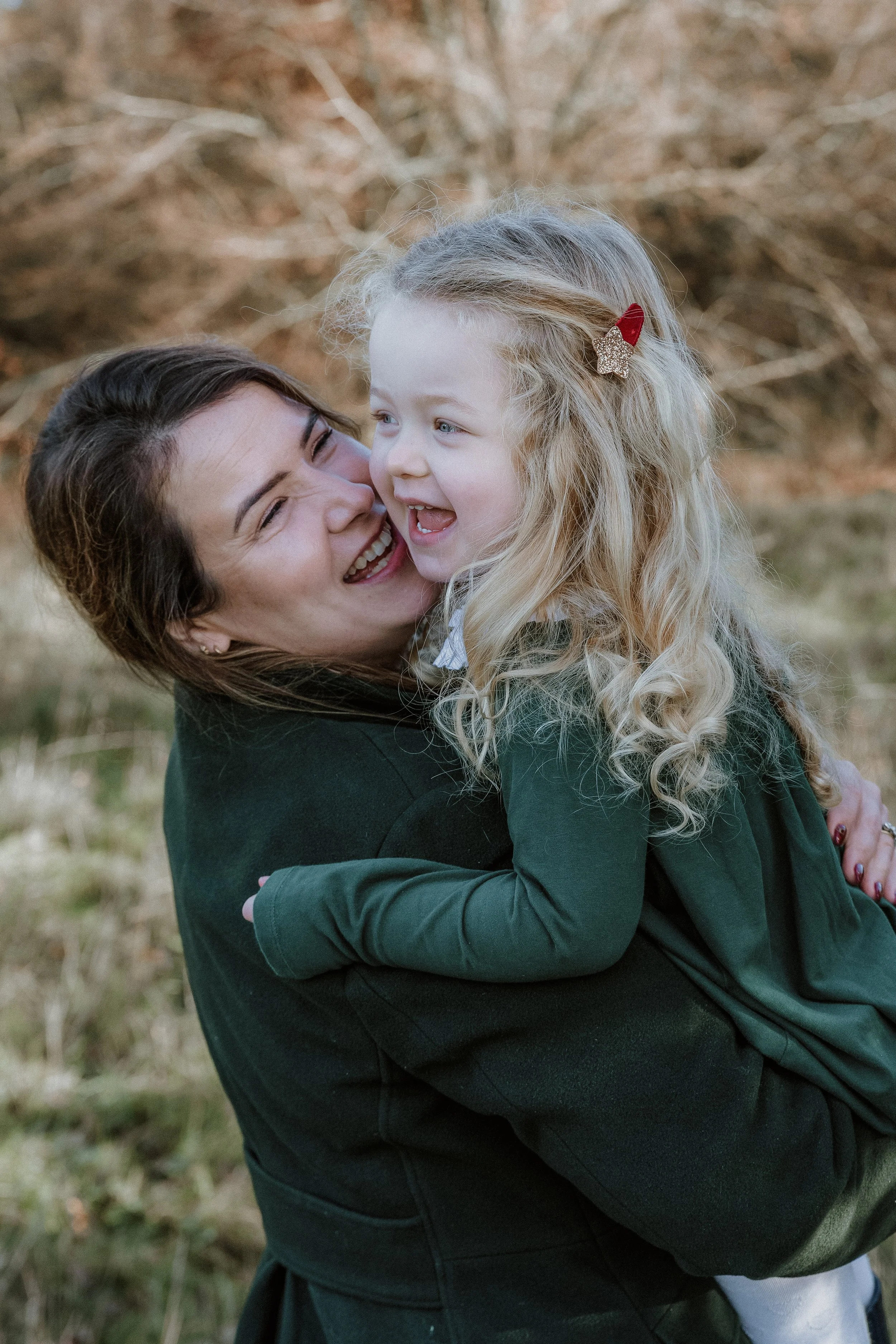 A woman holding a young girl outdoors, both smiling and laughing, with trees in the background.