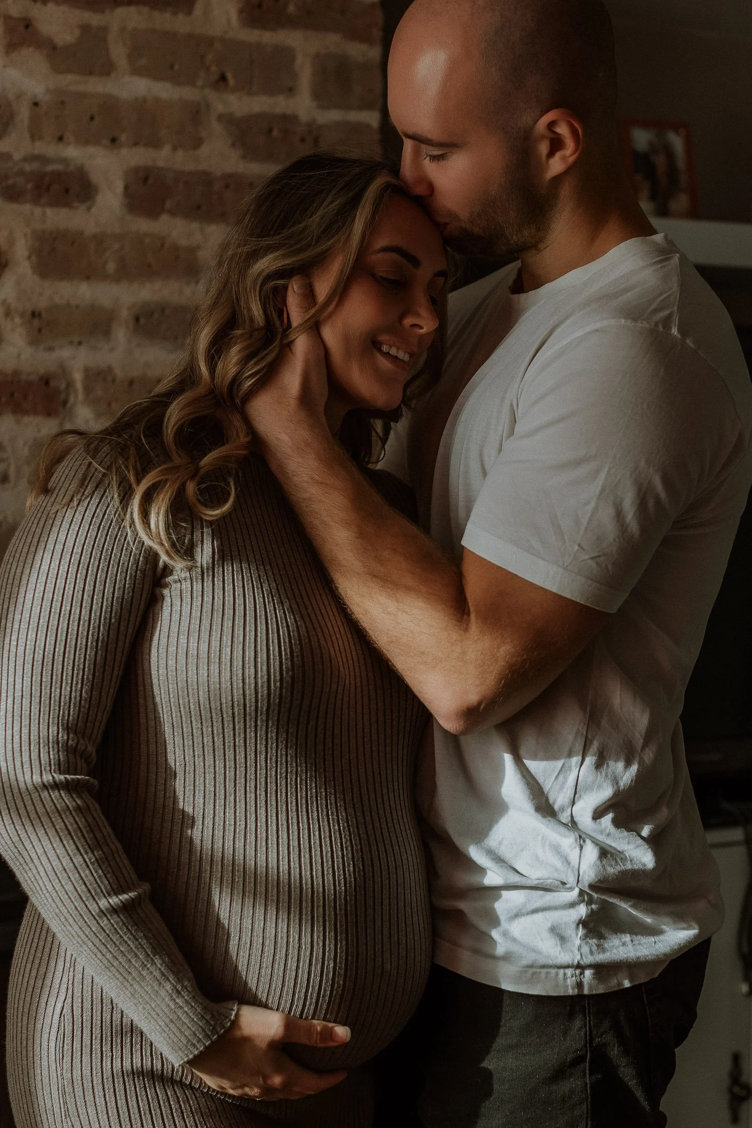 A pregnant woman with curly hair and a striped dress being embraced by a man with a shaved head and a white shirt, in a cozy indoor setting with a brick wall background.