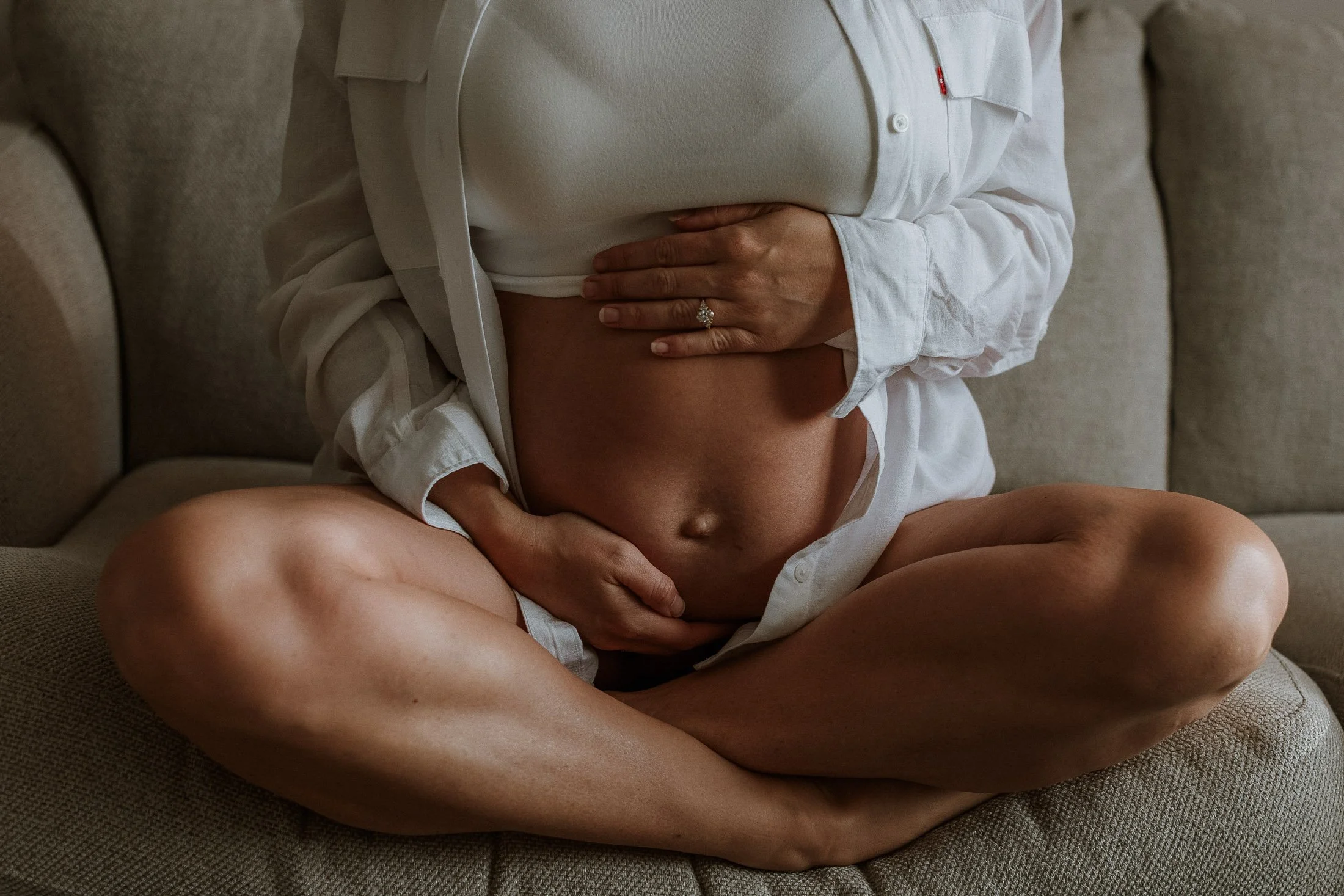 A pregnant woman sitting cross-legged on a sofa, lifting her white shirt to reveal her belly, with her hands gently resting on her stomach, wearing a wedding ring.