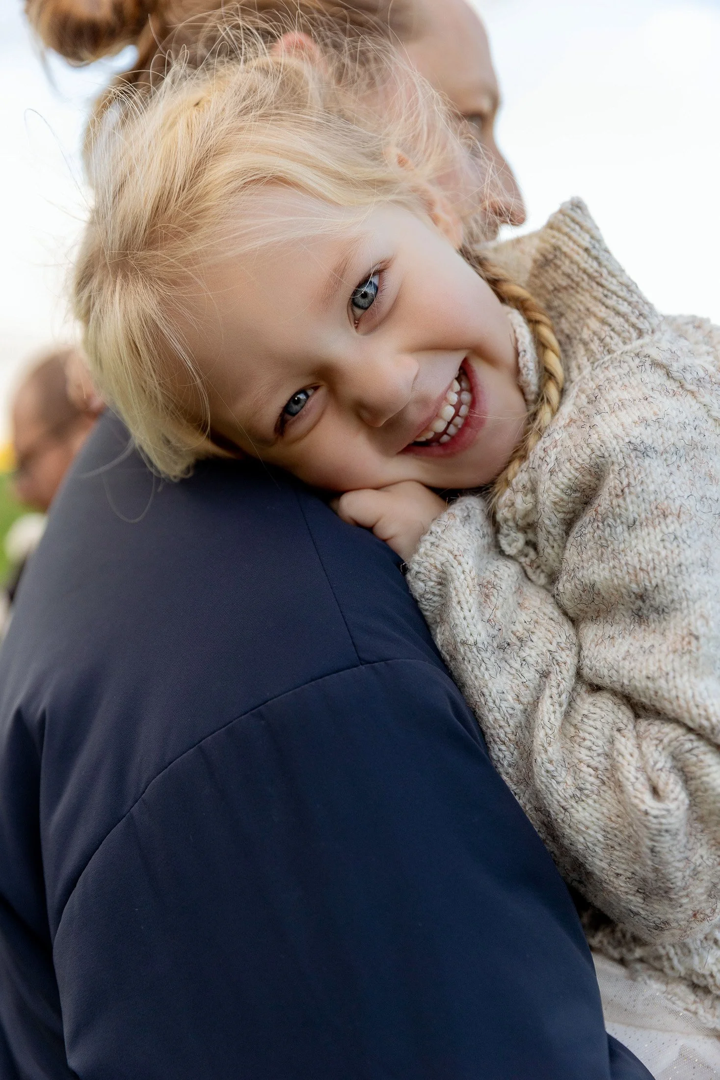 A young girl with blonde hair and blue eyes smiling while resting her head on someone's shoulder.