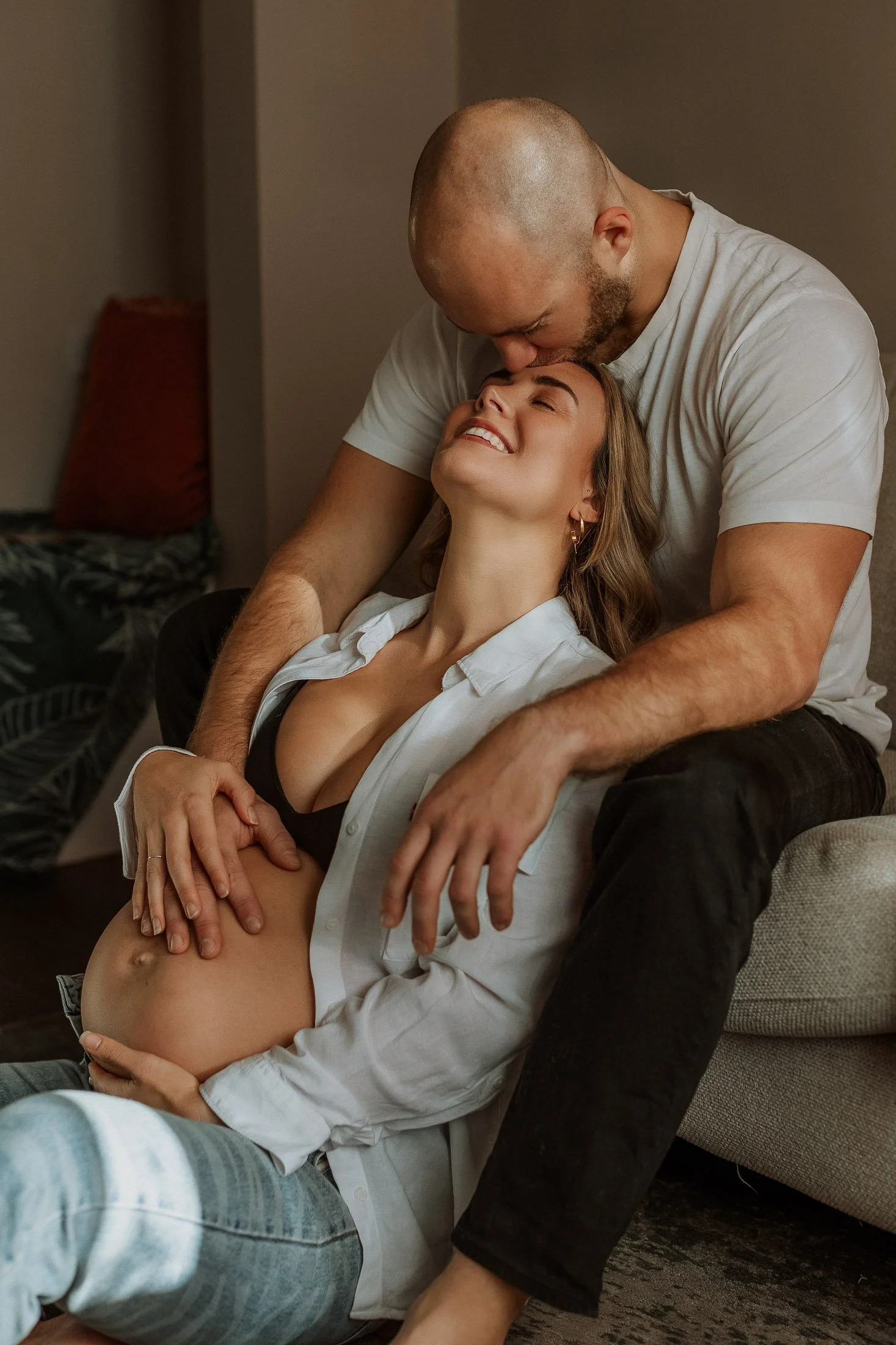 A pregnant woman smiling while sitting on the floor with her head tilted back, being embraced and kissed on the forehead by a man who is sitting behind her on a couch.