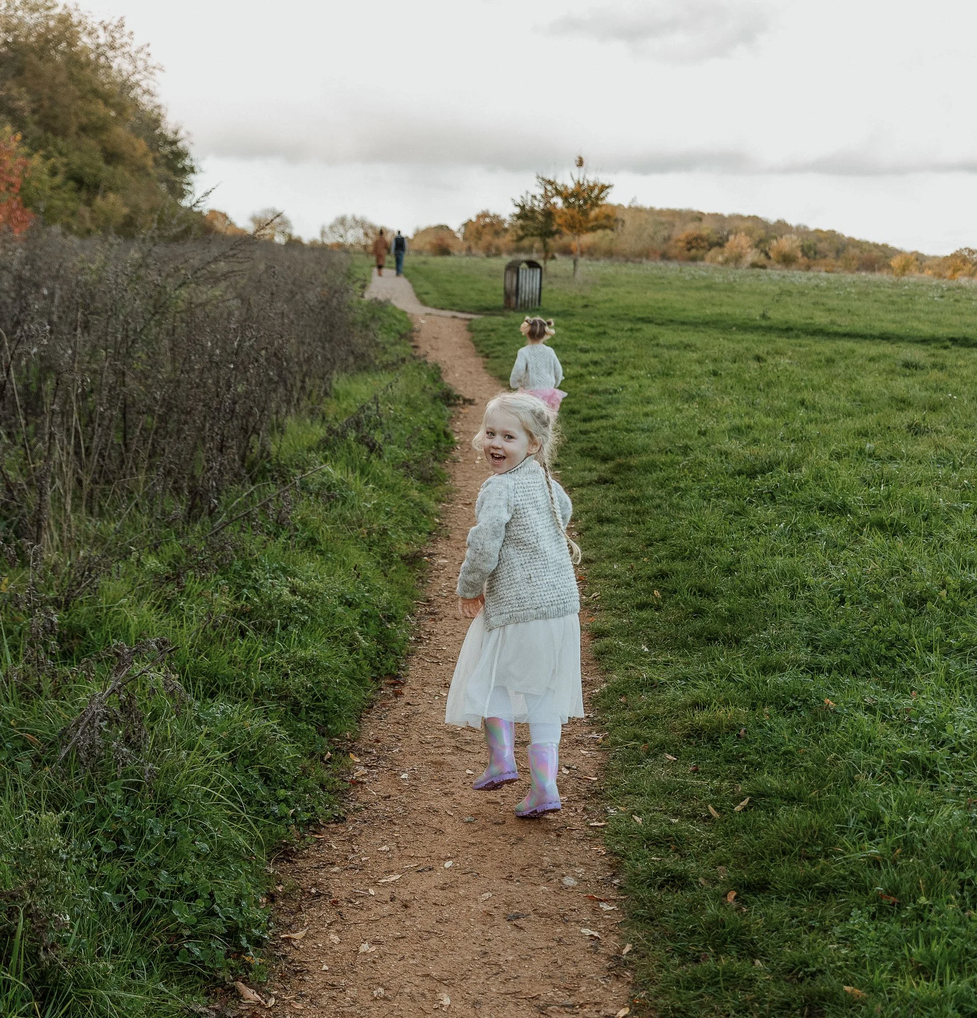 A young girl wearing a white dress and a gray sweater, with iridescent rain boots, smiling and looking back as she walks along a dirt path in a grassy park, with other children and adults in the background.