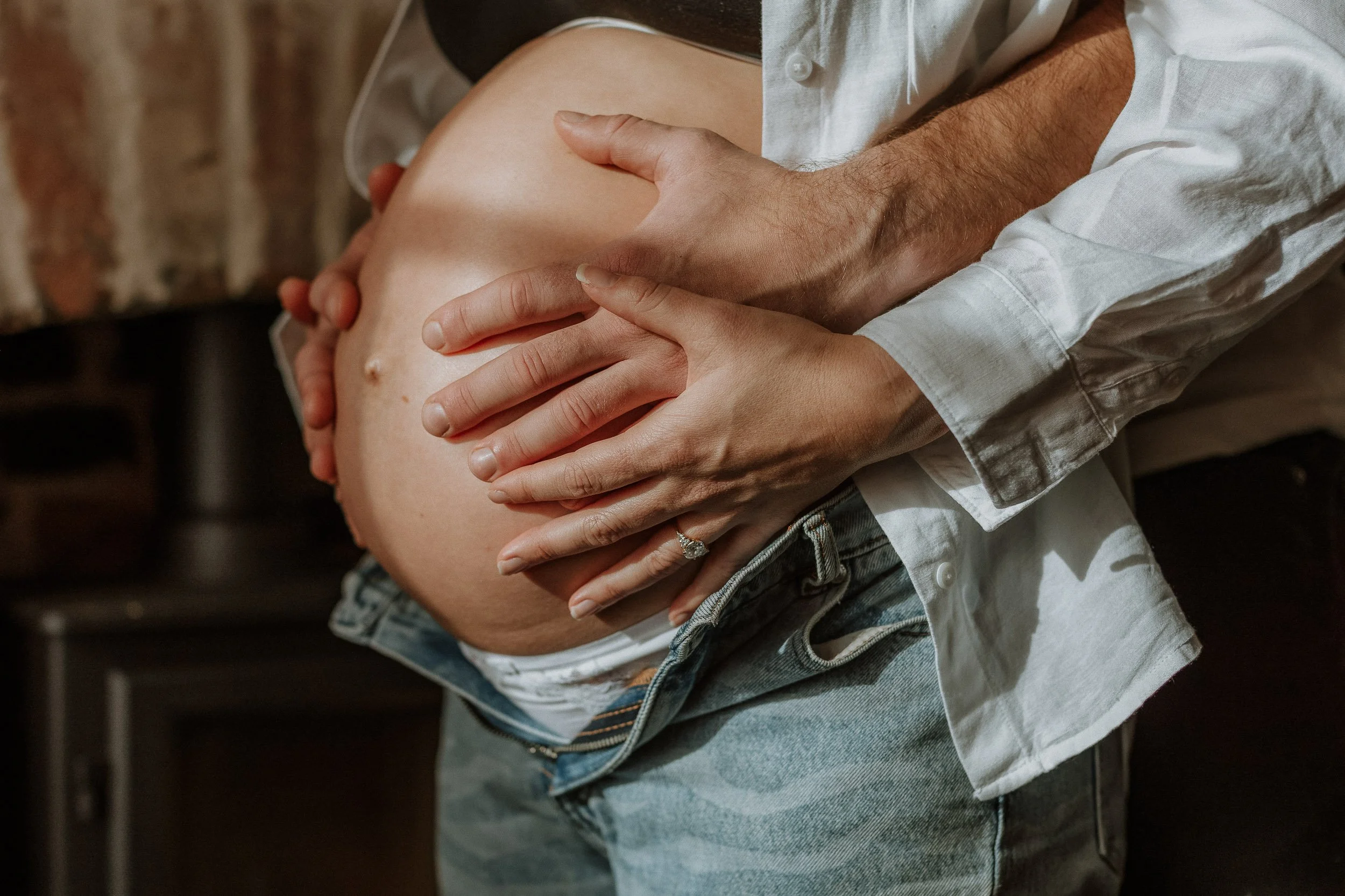 A man gently holding a pregnant woman's belly with both hands; she is wearing jeans and an open white shirt, and he is wearing a white shirt.