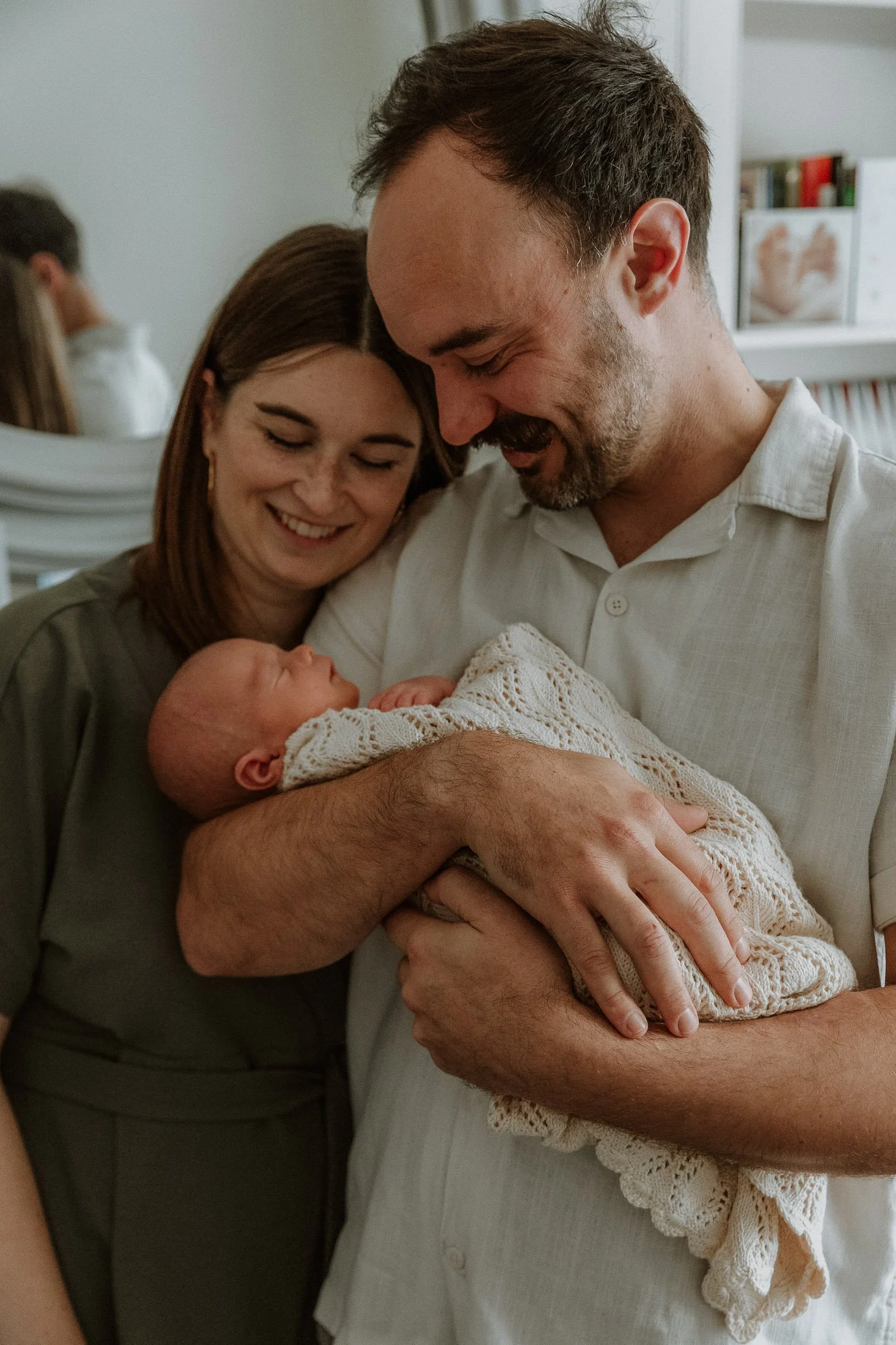 A man and woman with a newborn baby, smiling and looking at the baby, in a cozy indoor setting.