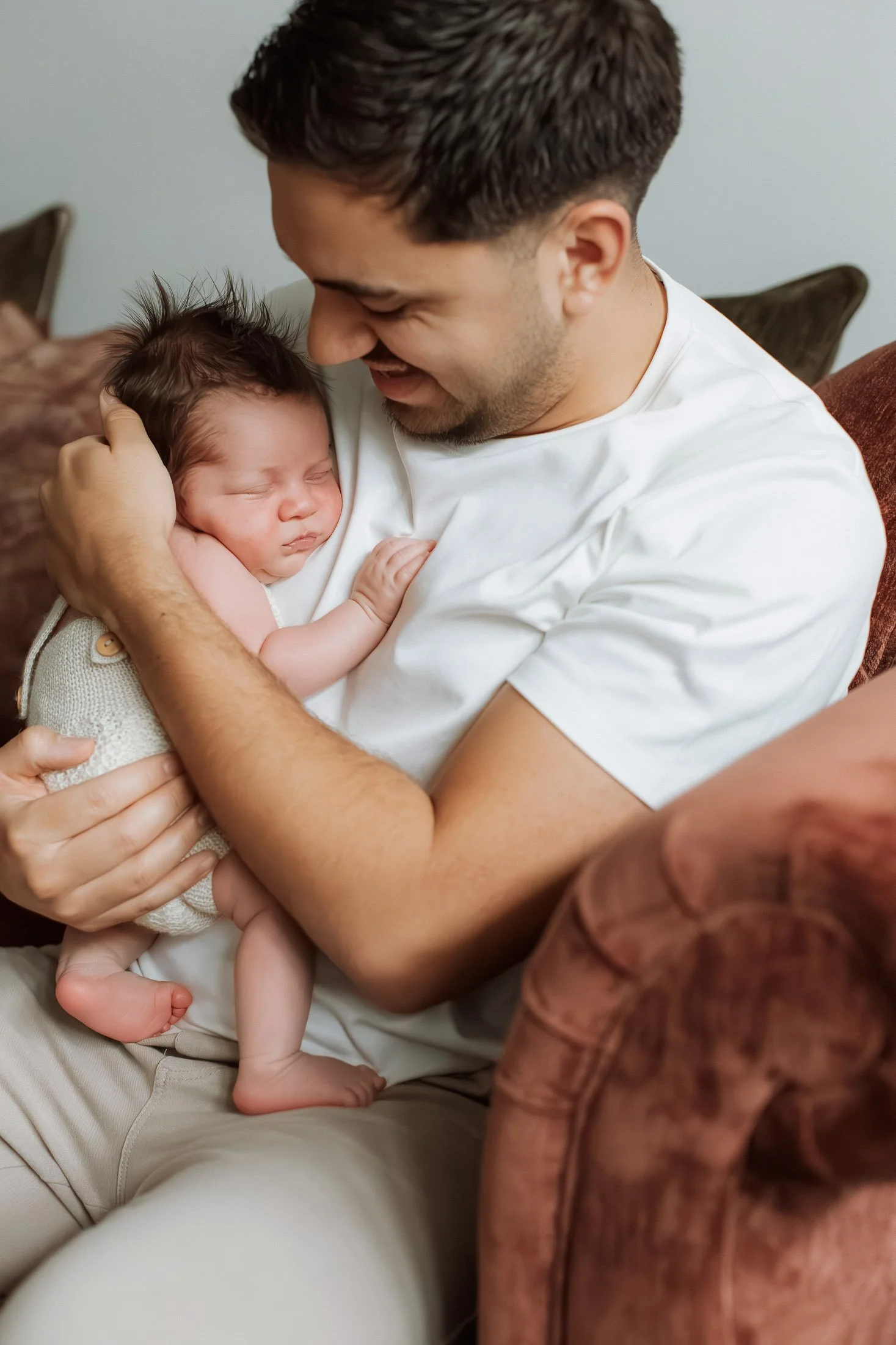 A man smiling and holding a sleeping baby on his chest while sitting on a sofa.