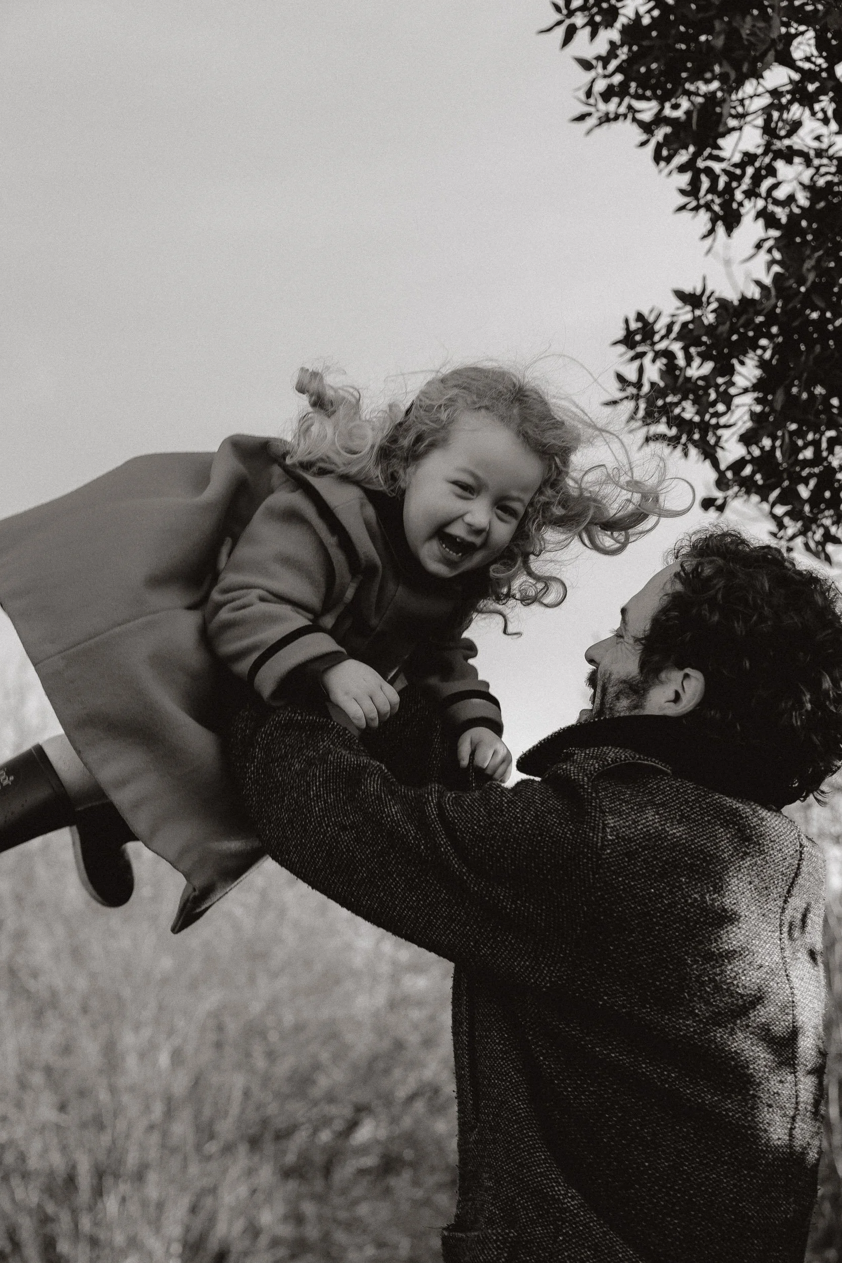 A man lifting a smiling young girl in the air outdoors, with trees in the background, in black and white.