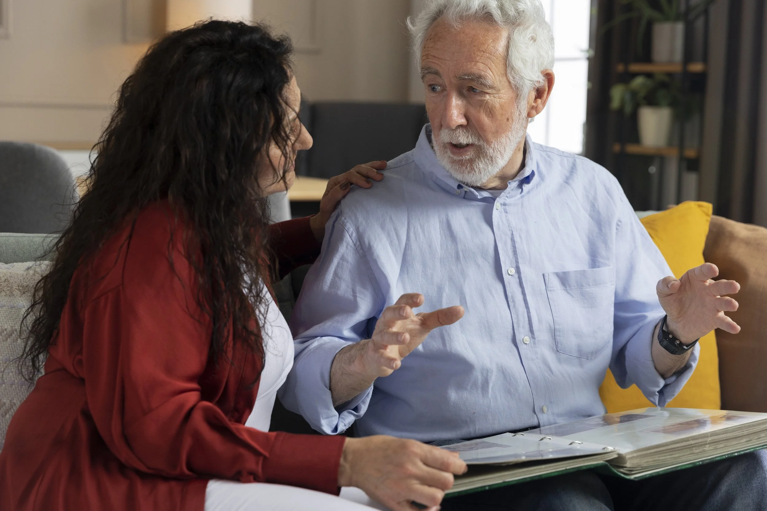 Deux personnes discutent, une femme à gauche et un homme à droite, dans un intérieur moderne, la femme porte un vêtement rouge et l'homme porte une chemise bleue, ils sont assis à une table avec un classeur ou un document.