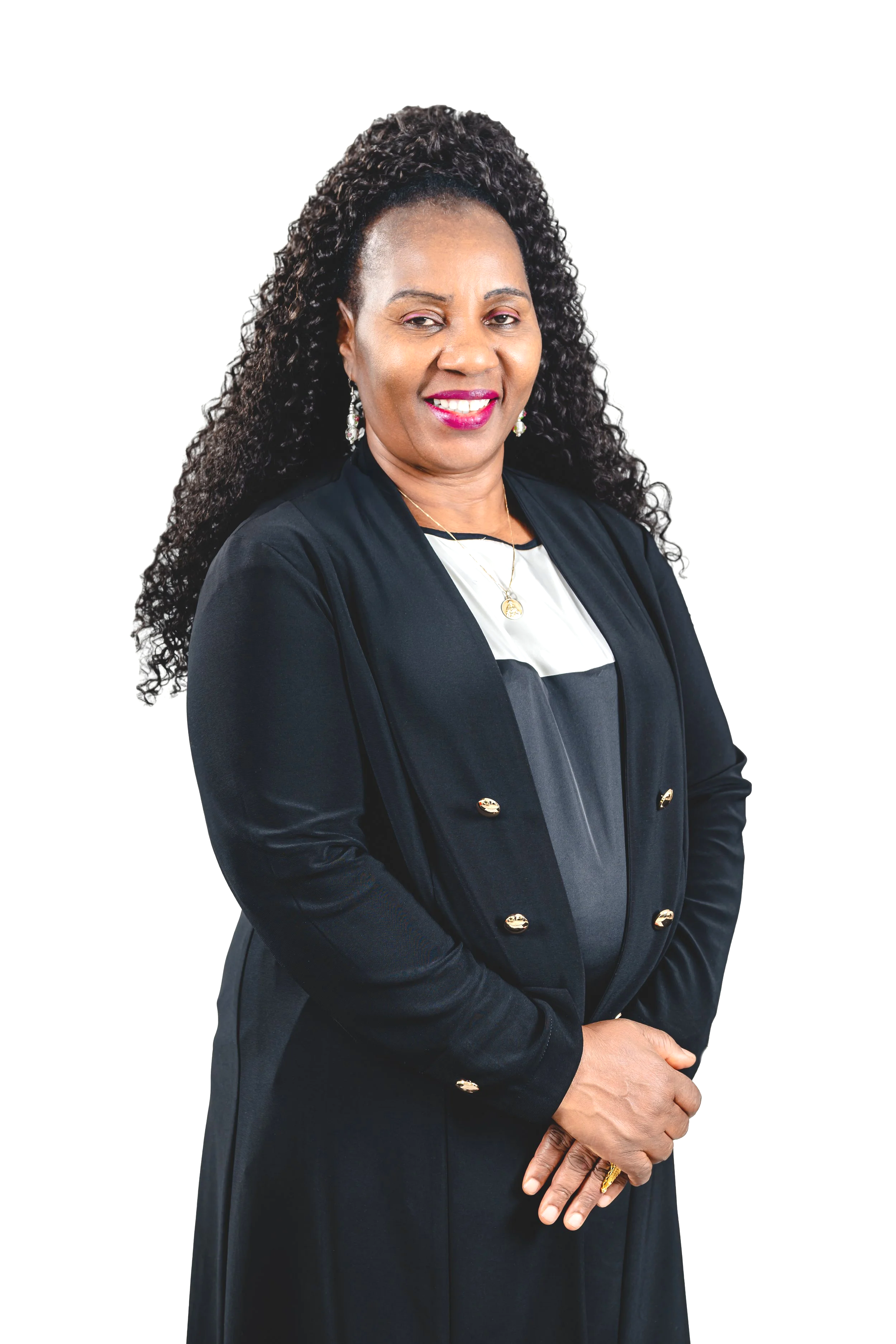 Une femme d'âge moyen avec de longs cheveux bouclés porte une robe noire et une veste noire orné de boutons dorés. Elle porte également des boucles d'oreilles pendantes, un collier, et une bague dorée. Elle sourit légèrement et pose les bras croisés.