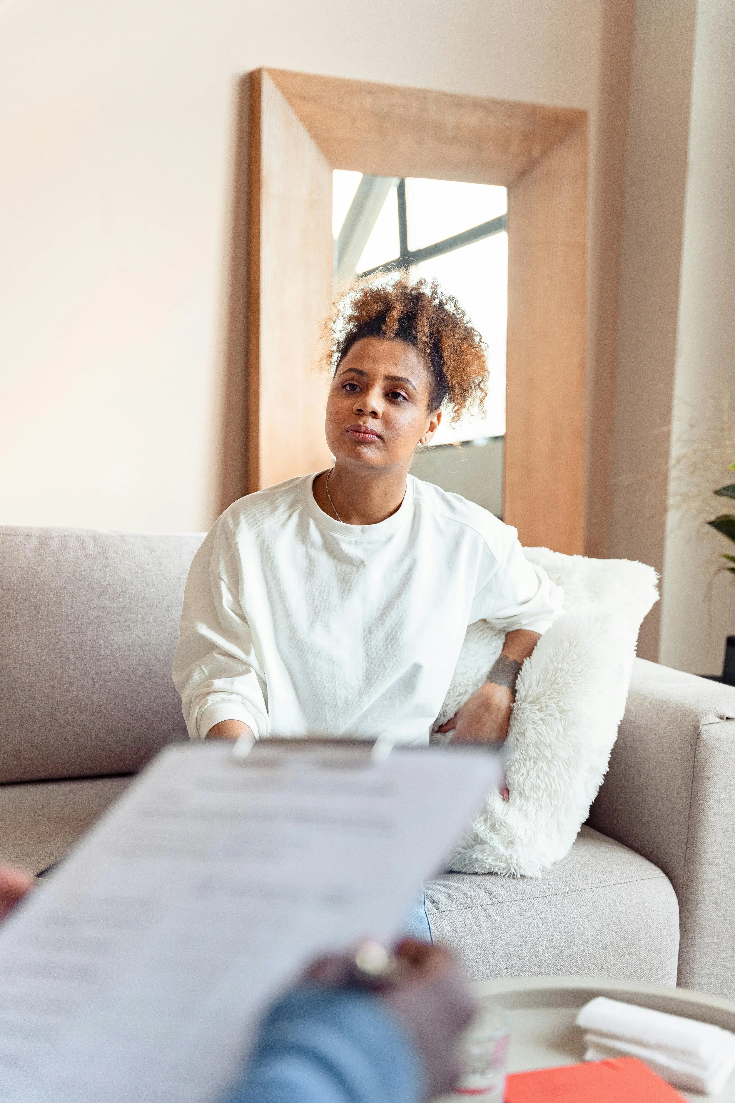 Une jeune femme assise sur un canapé en tissu beige, regardant pensivement devant elle, avec un coussin blanc à ses côtés, dans un intérieur lumineux avec un miroir en bois derrière elle.
