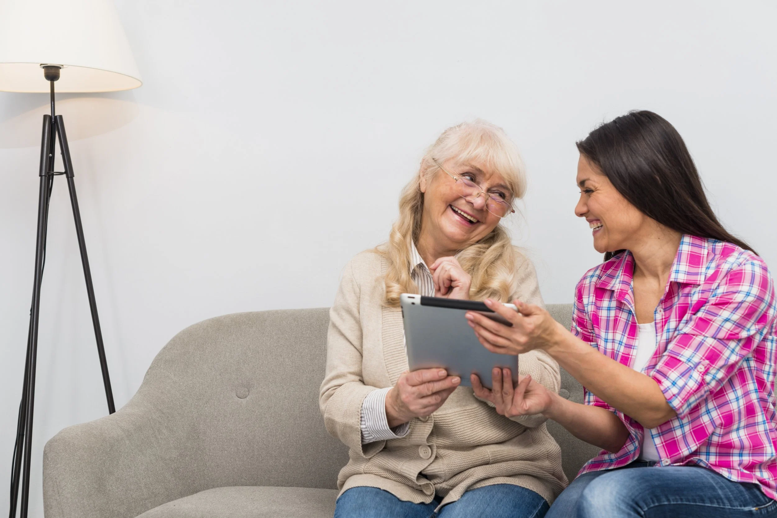 Deux femmes souriantes regardent une tablette, assises sur un canapé gris, dans un intérieur lumineux avec une lampe sur pied noire et blanche à gauche.