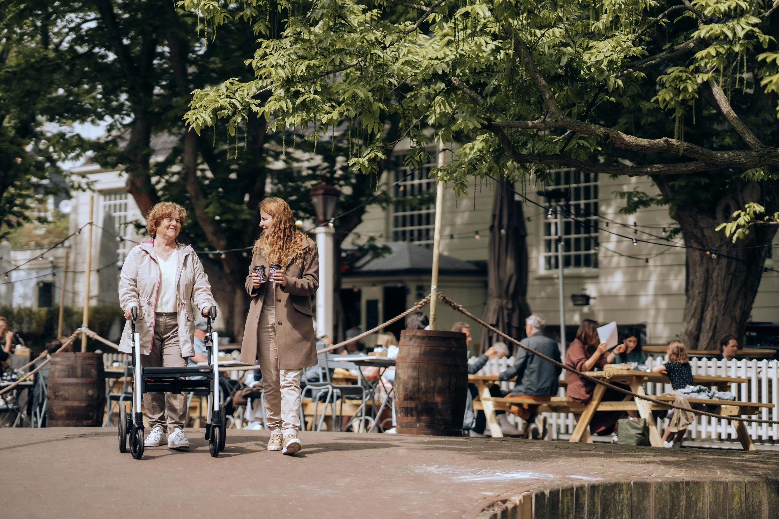Deux femmes âgées marchent dans une rue pavée, l'une d'elles pousse une marchette. Elles se sourient et tiennent des tasses à café. Il y a des tables d'extérieur avec des gens assis, et de grands arbres avec des feuilles vertes au-dessus.