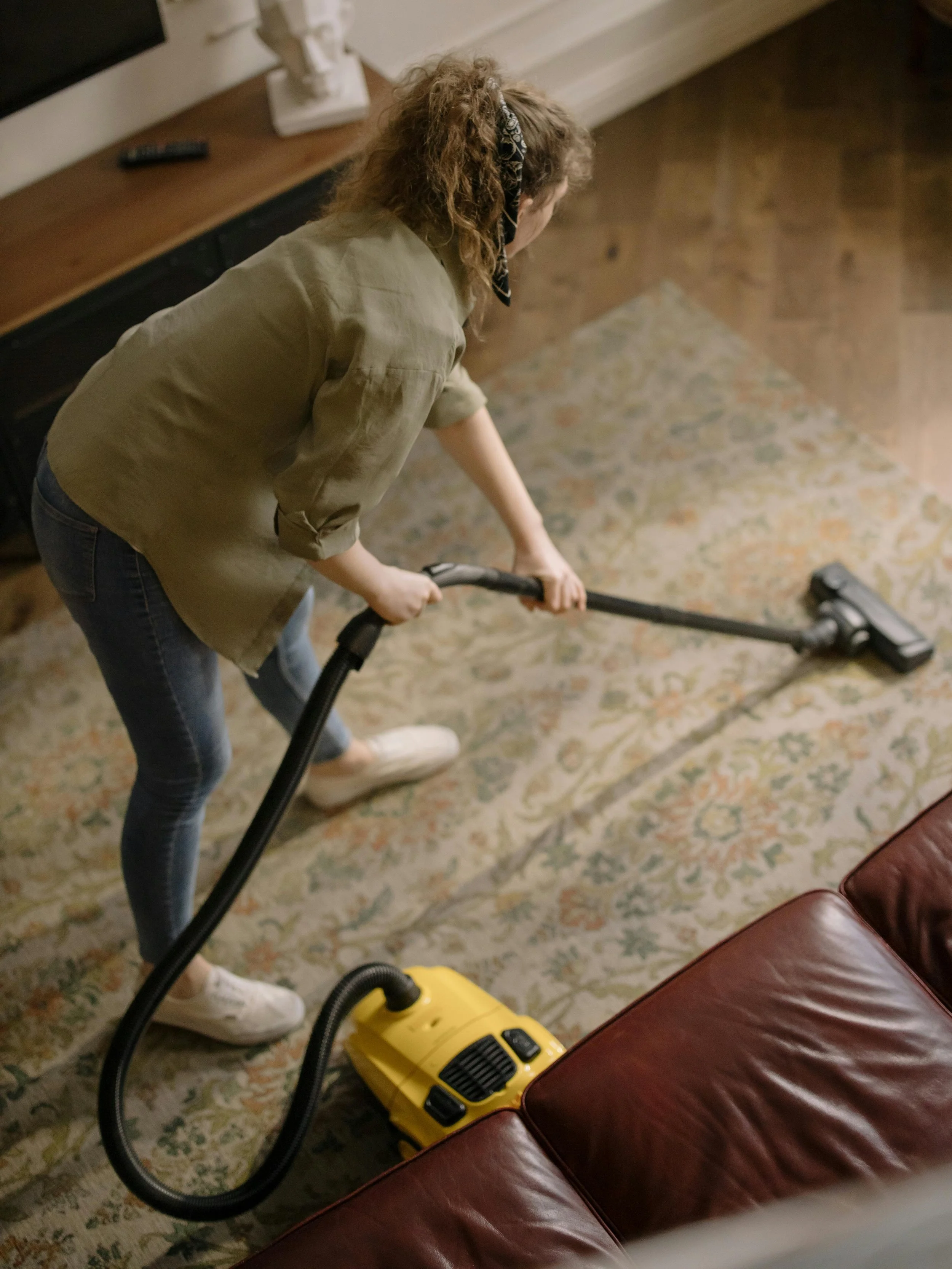 Femme avec cheveux bouclés et foulard nettoyant un tapis avec un aspirateur jaune dans un salon avec tapis en motif et fauteuil en cuir marron.