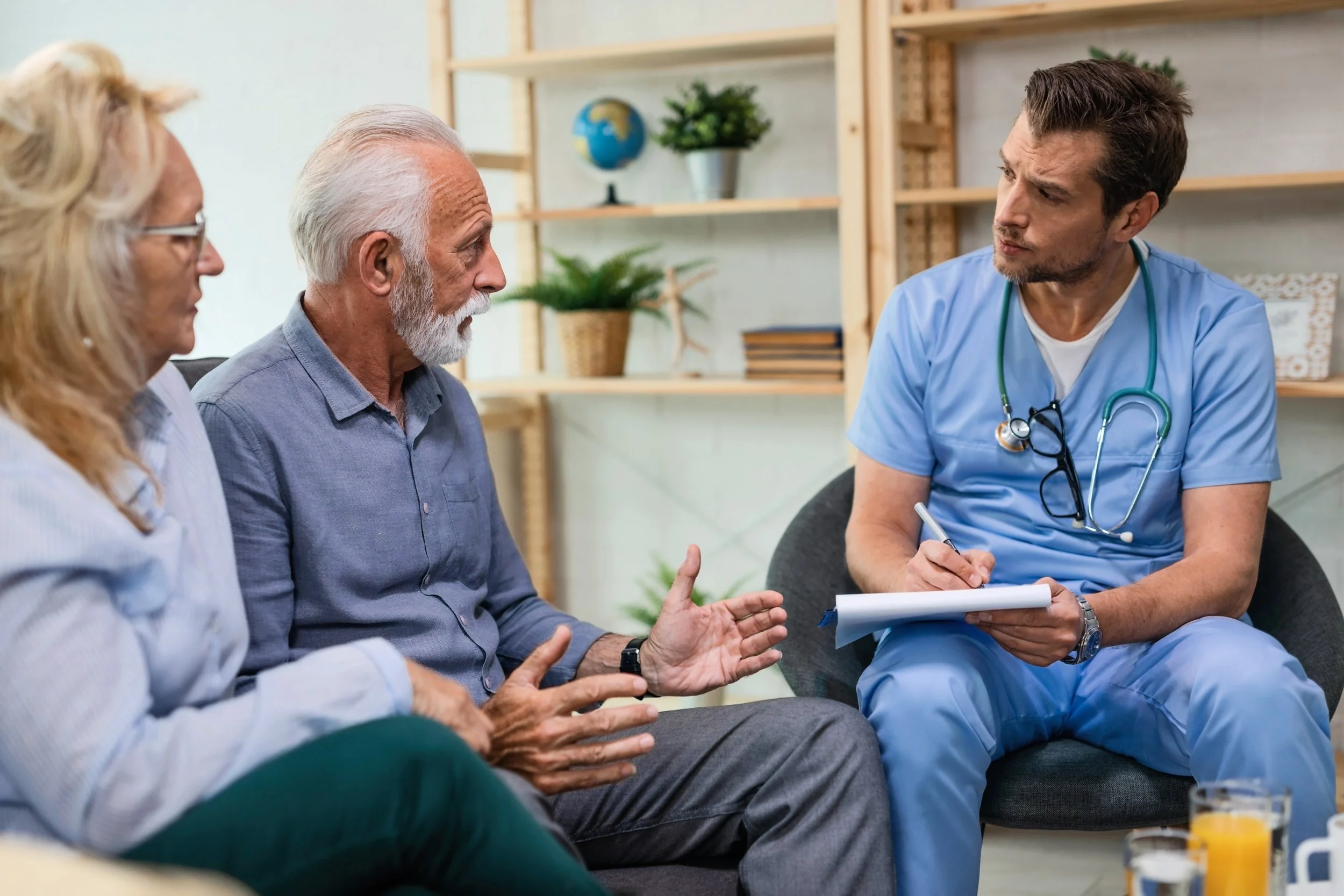 Un groupe de personnes, comprenant un médecin en tenue médicale bleue avec stéthoscope, discute dans un cadre intérieur avec des étagères en bois et des plantes décoratives.