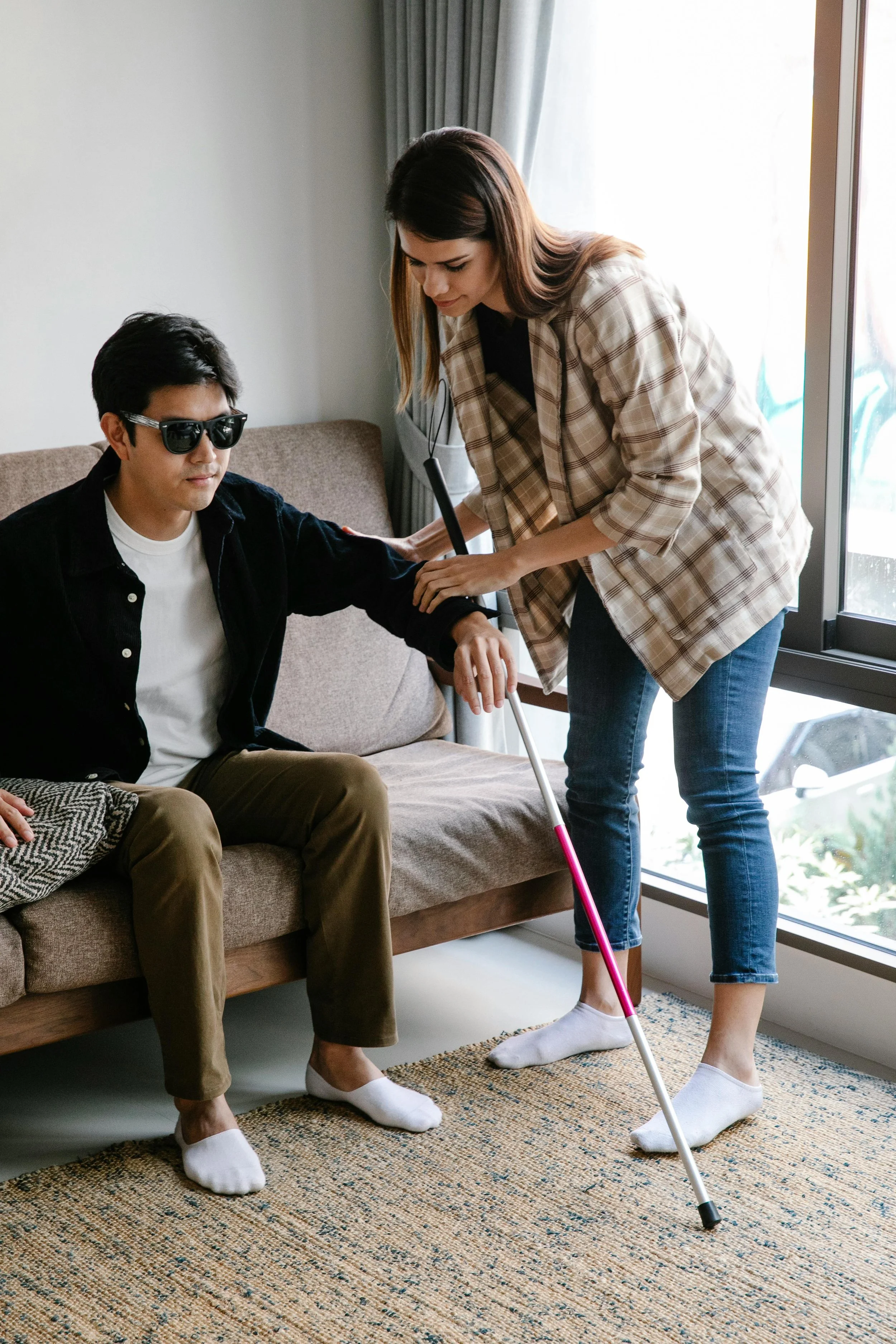 Une femme aide un homme malvoyant avec une canne dans un salon lumineux, ils portent tous deux des chaussettes blanches.