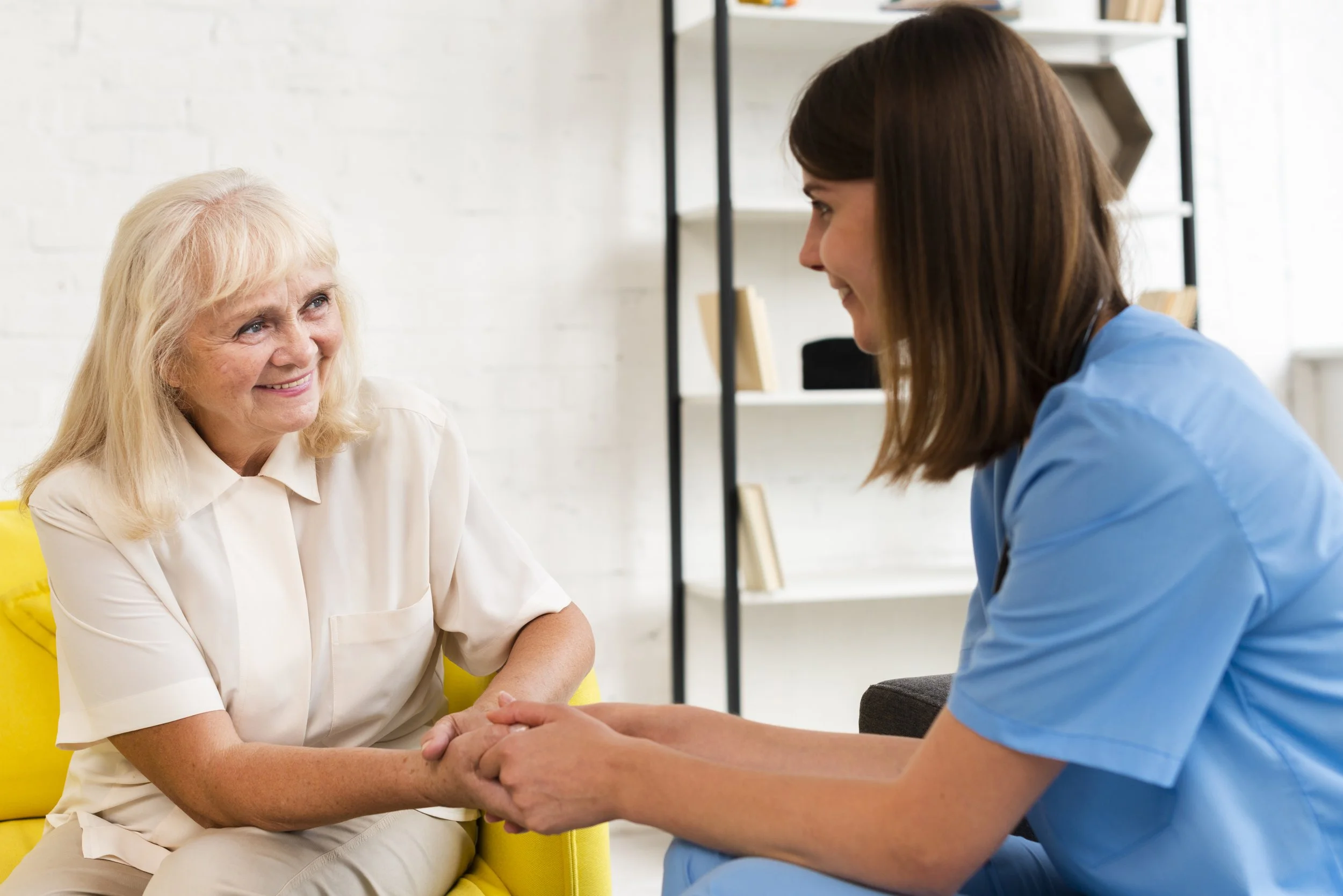 Une femme âgée souriante bavarde avec une jeune femme en uniforme médical, main dans la main, dans un environnement intérieur moderne.