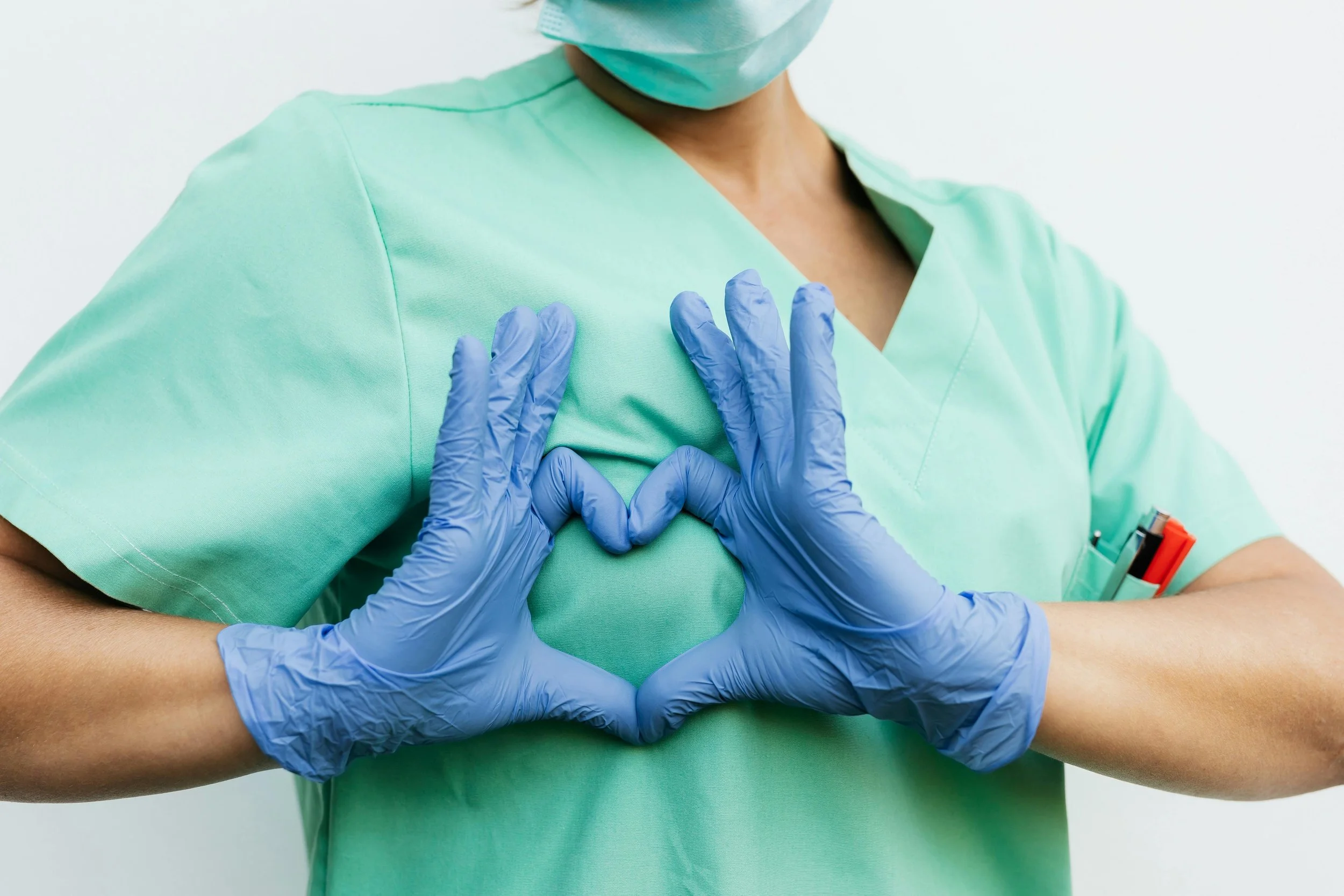Une personne en blouse médicale et masque chirurgical, portant des gants en latex bleus, fait le geste de l'amour avec ses mains sur sa poitrine.