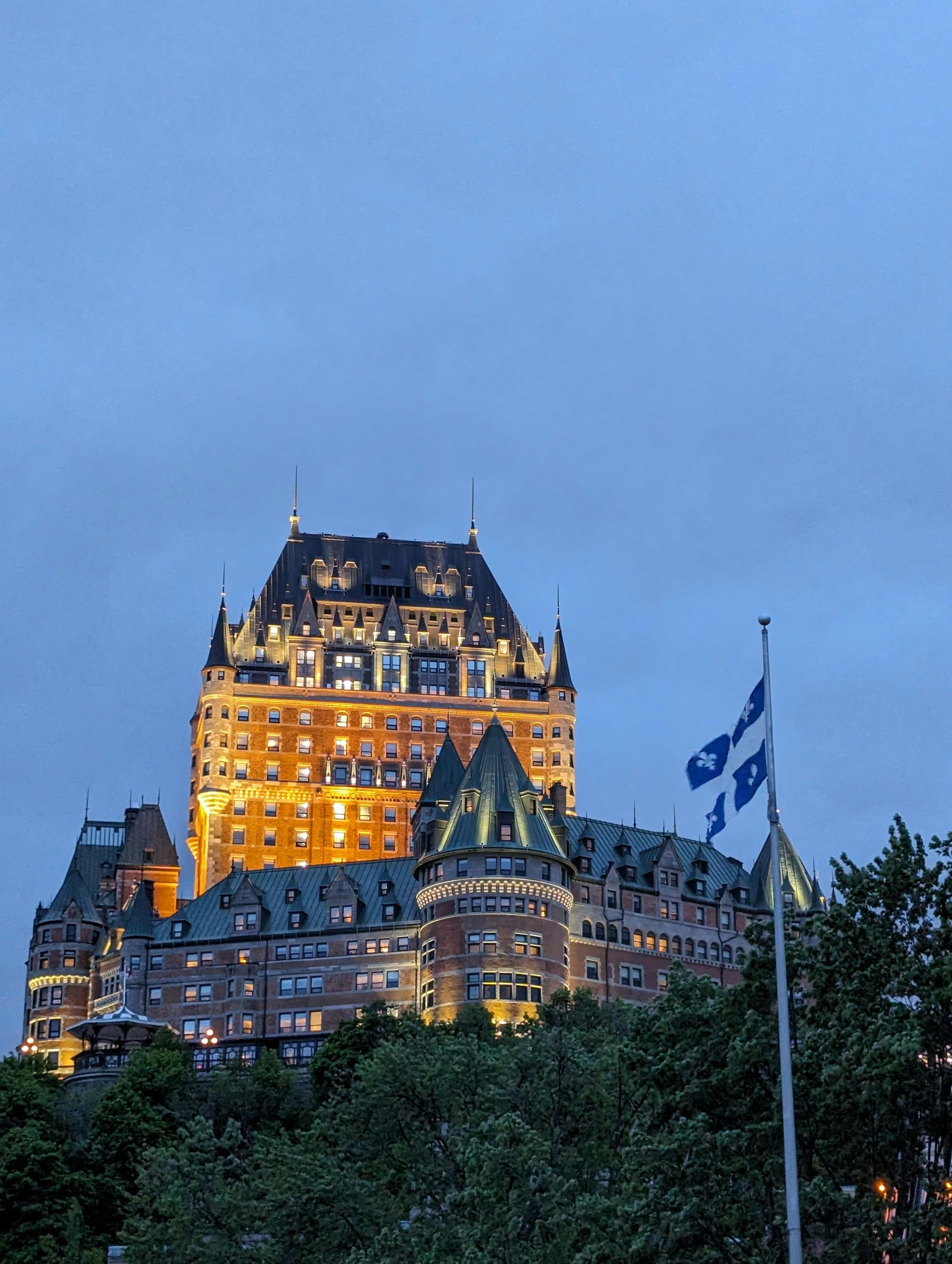 Un grand château historique illuminé la nuit avec une tour centrale et plusieurs toits en pente, entouré d'arbres et un drapeau québécois flottant à côté.
