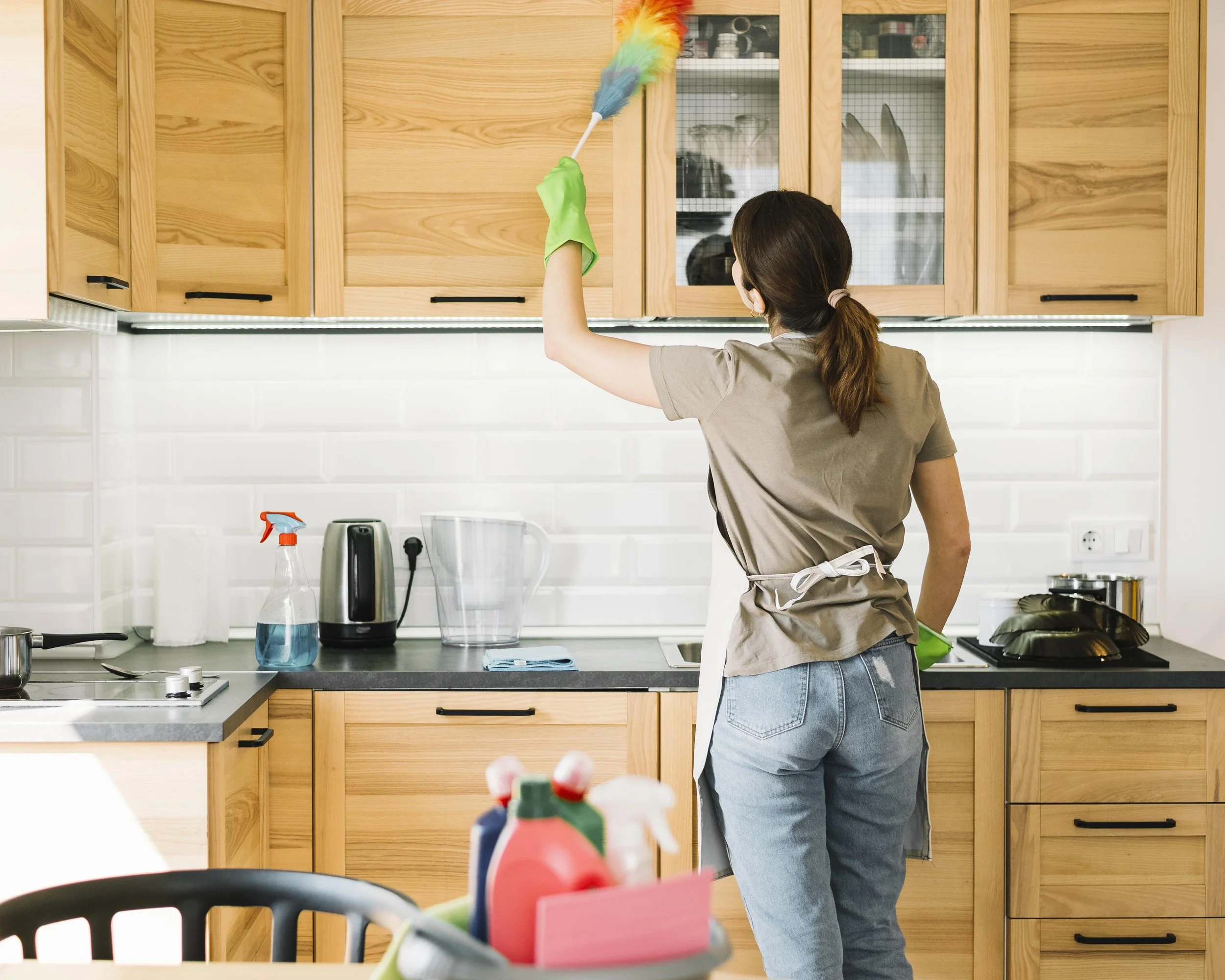 Une femme nettoie des armoires de cuisine en utilisant une brosse à manche long. La cuisine a des meubles en bois clair et un plan de travail gris. Il y a des objets de nettoyage et des appareils électriques sur le plan de travail.