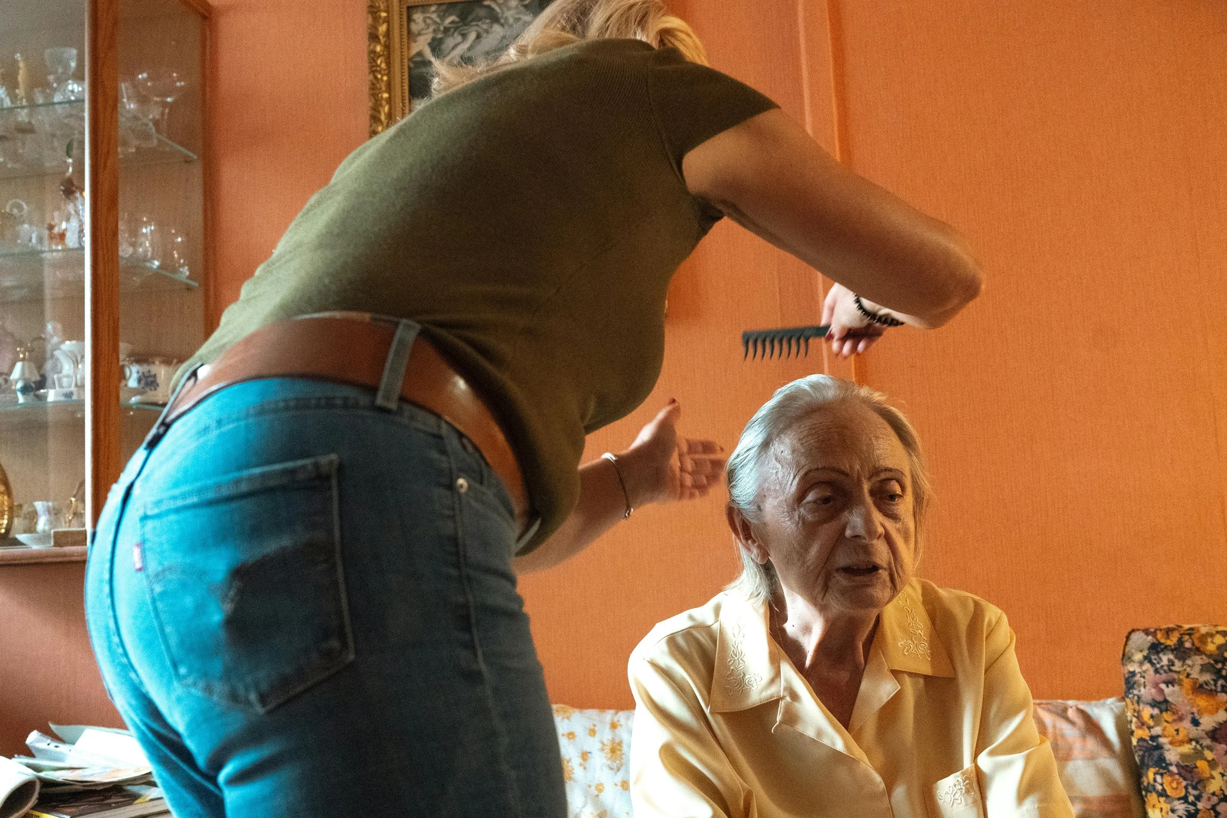 Une femme âgée assise, une autre femme debout lui parlant, dans une pièce à décor orange avec une vitrine en bois contenant de la vaisselle, et un tableau accroché au mur.