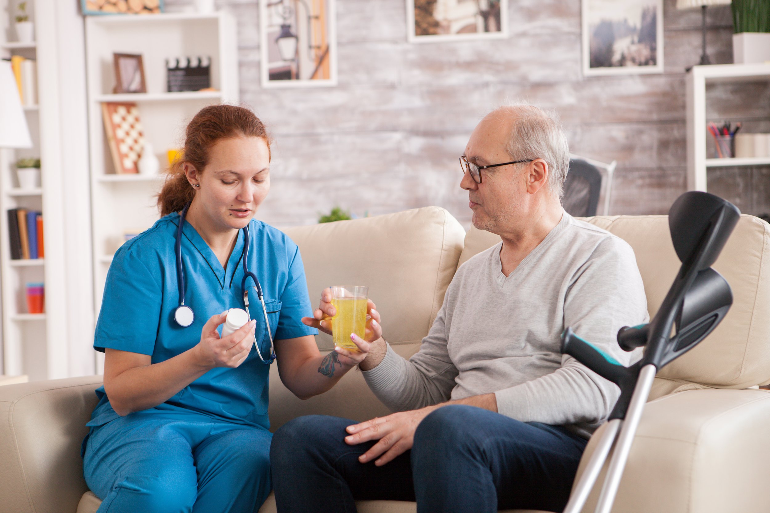 Une infirmière en uniforme bleu remet des médicaments à un homme âgé en fauteuil roulant. L'homme tient un verre de boisson. La scène se déroule dans un salon, avec des étagères contenant des livres et des décorations en arrière-plan.