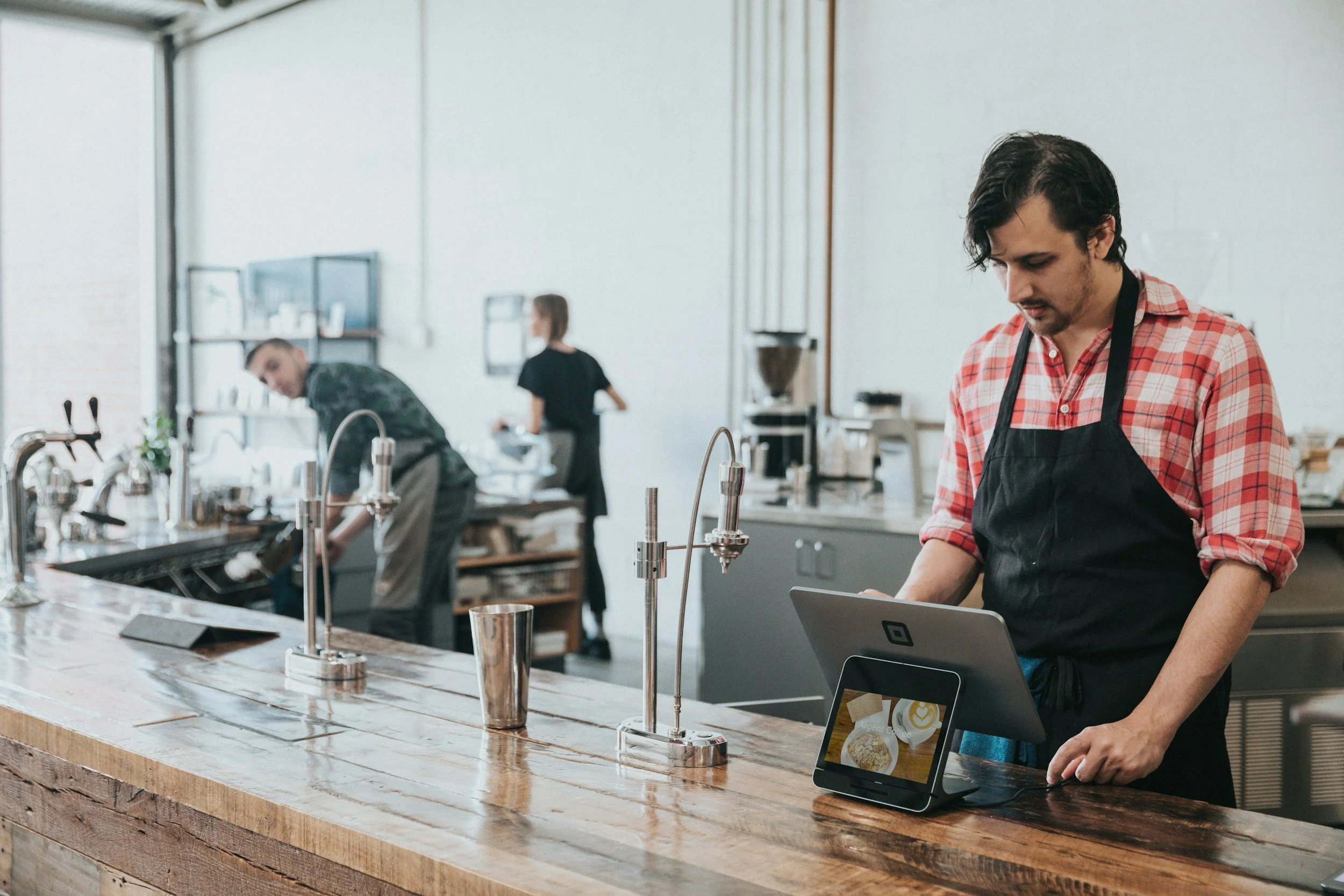 Barista working at the counter with a point of sale system in a modern cafe, with two other staff members in the background cleaning and preparing in a bright, minimalist space.