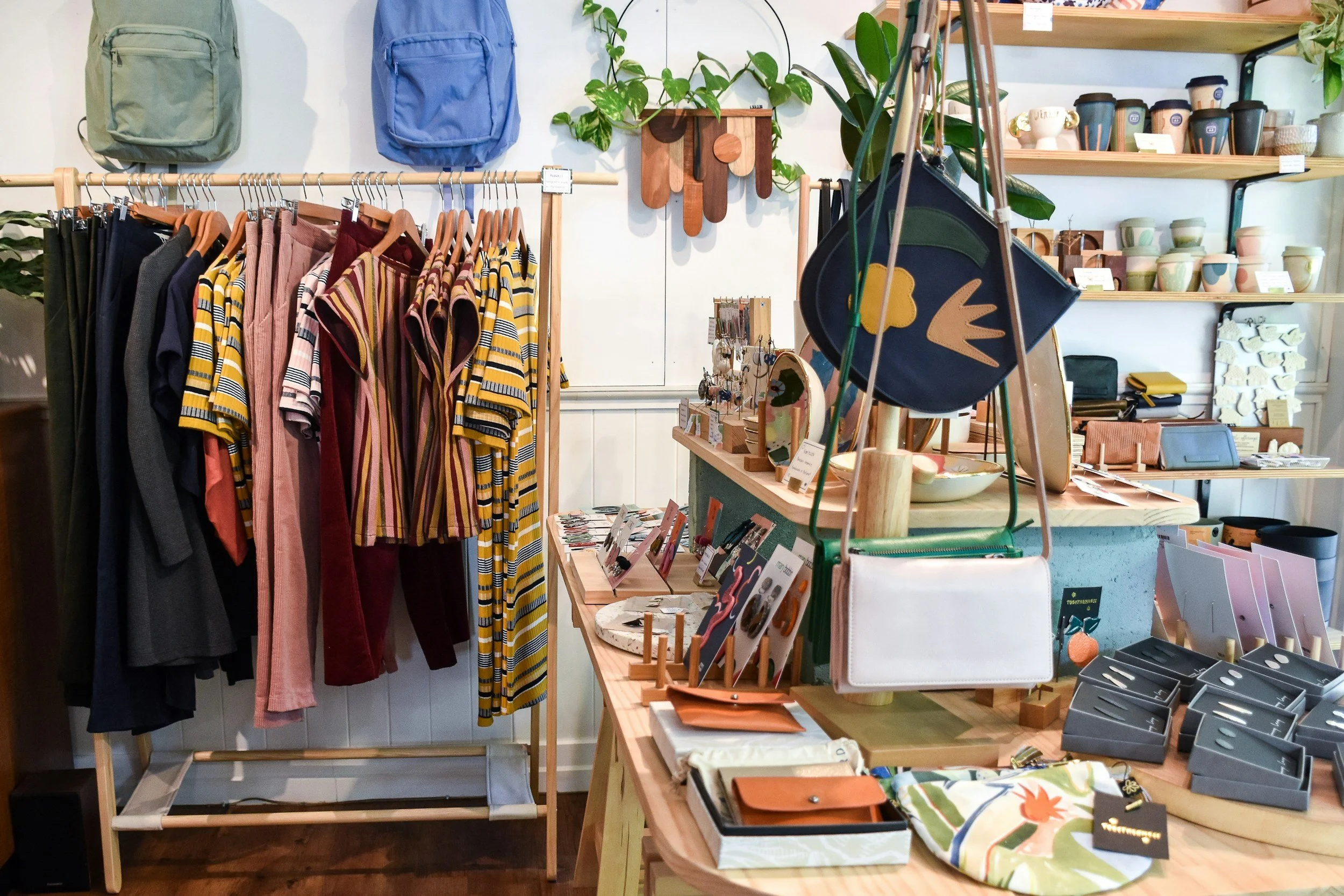 Interior of a retail store with clothing for sale on a wooden rack, including shirts and pants, along with accessories like bags, jewelry, and home goods on shelves and tables, all arranged in a cozy and stylish manner.