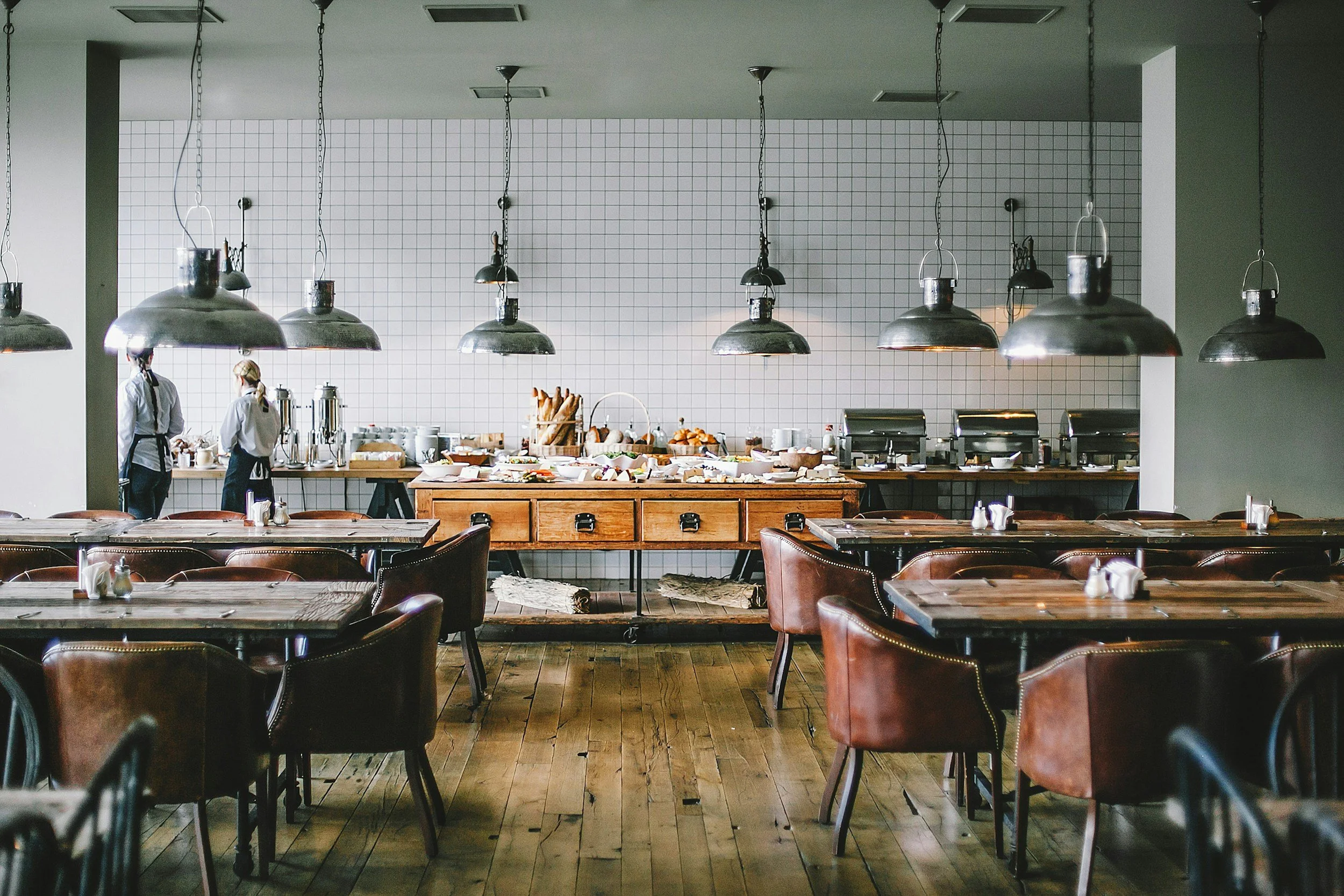 Empty restaurant dining area with dark wooden tables and leather chairs, industrial pendant lights hanging from the ceiling, and a buffet station with various breakfast foods in the background, including bread, cereals, and beverages, with two staff members attending in the kitchen area.