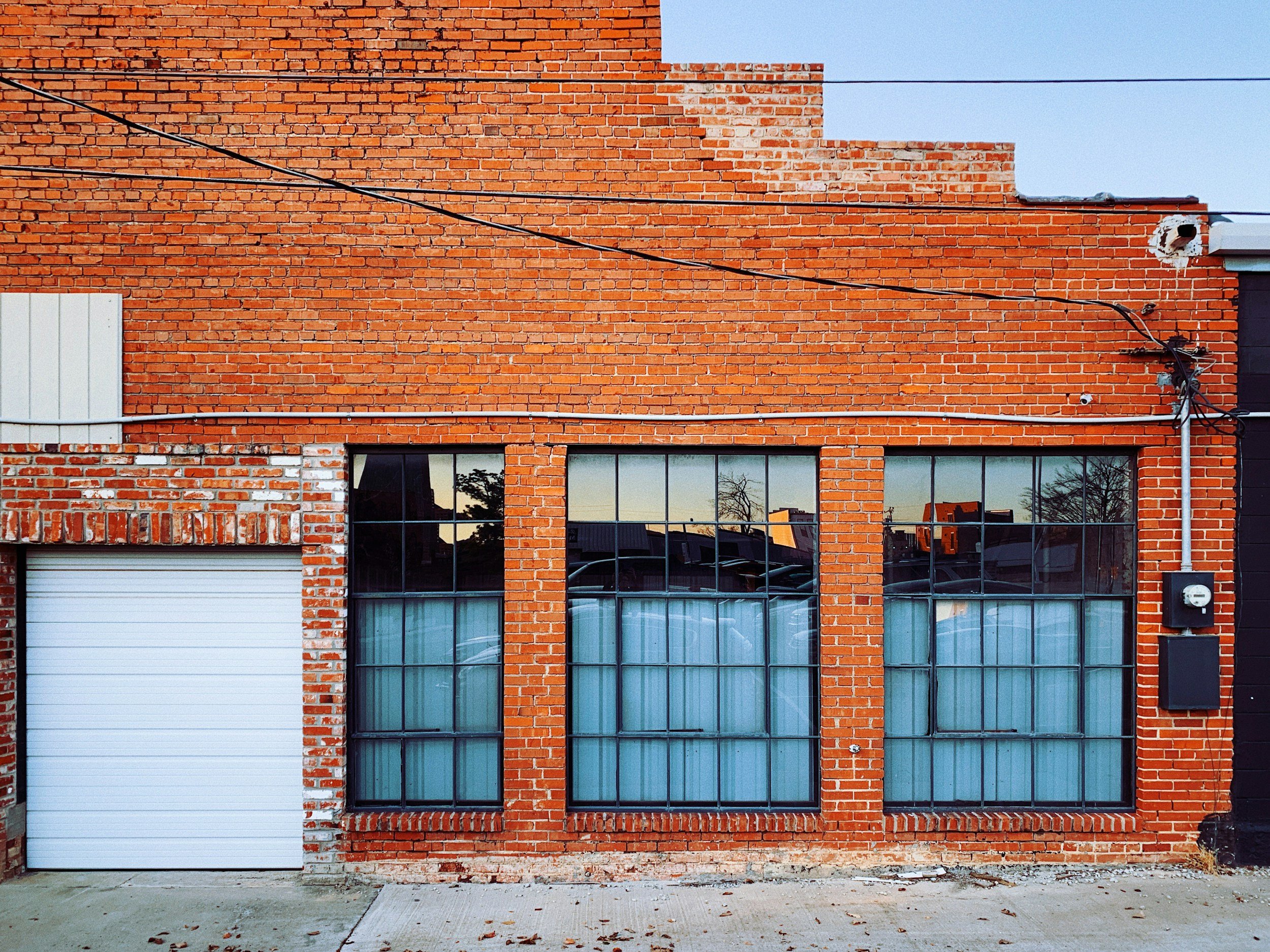 Red brick building with three large windows with black frames, one garage door on the left, and electrical wires overhead. The building shows signs of weathering, and there are reflections of other buildings and trees in the windows.