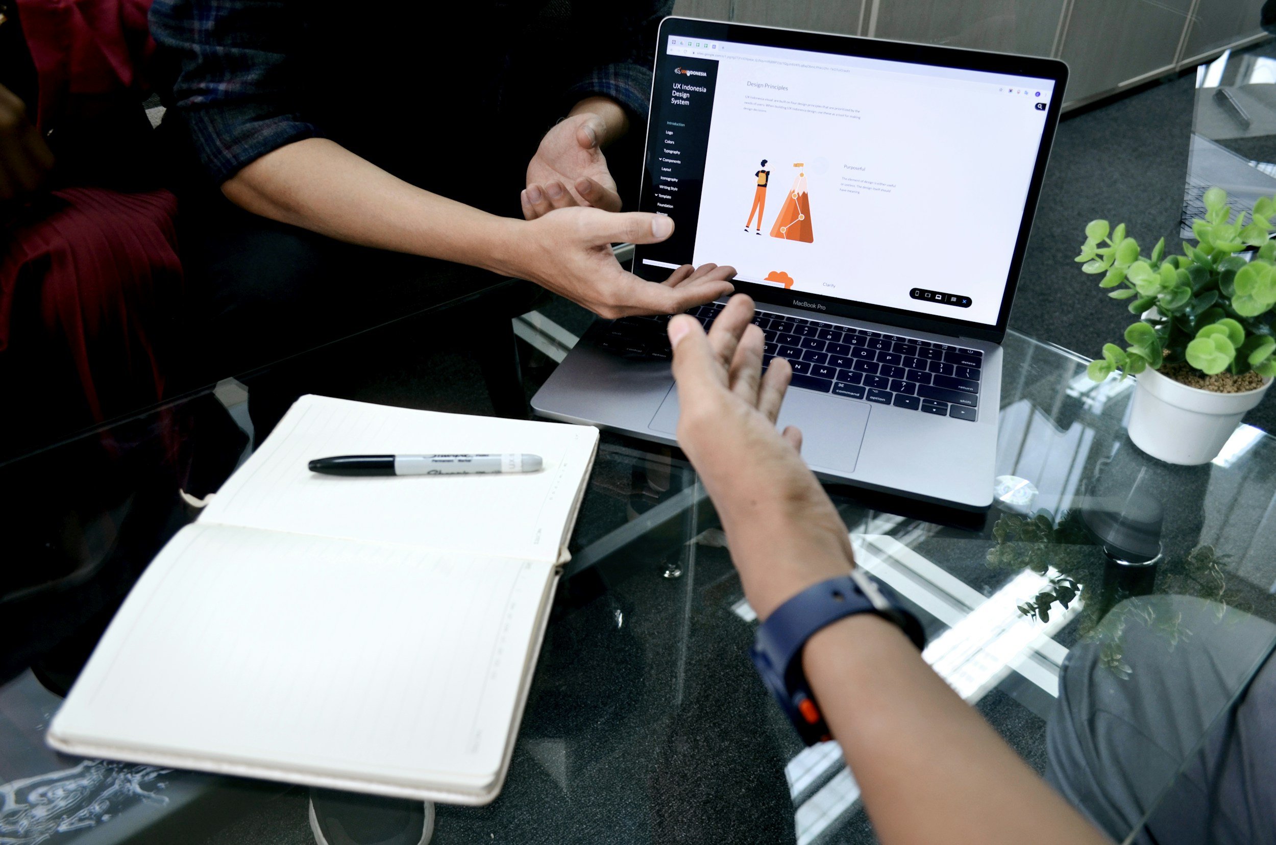 Two people discussing a design on a laptop screen, with a notebook and pen on the glass table, and a potted plant nearby.