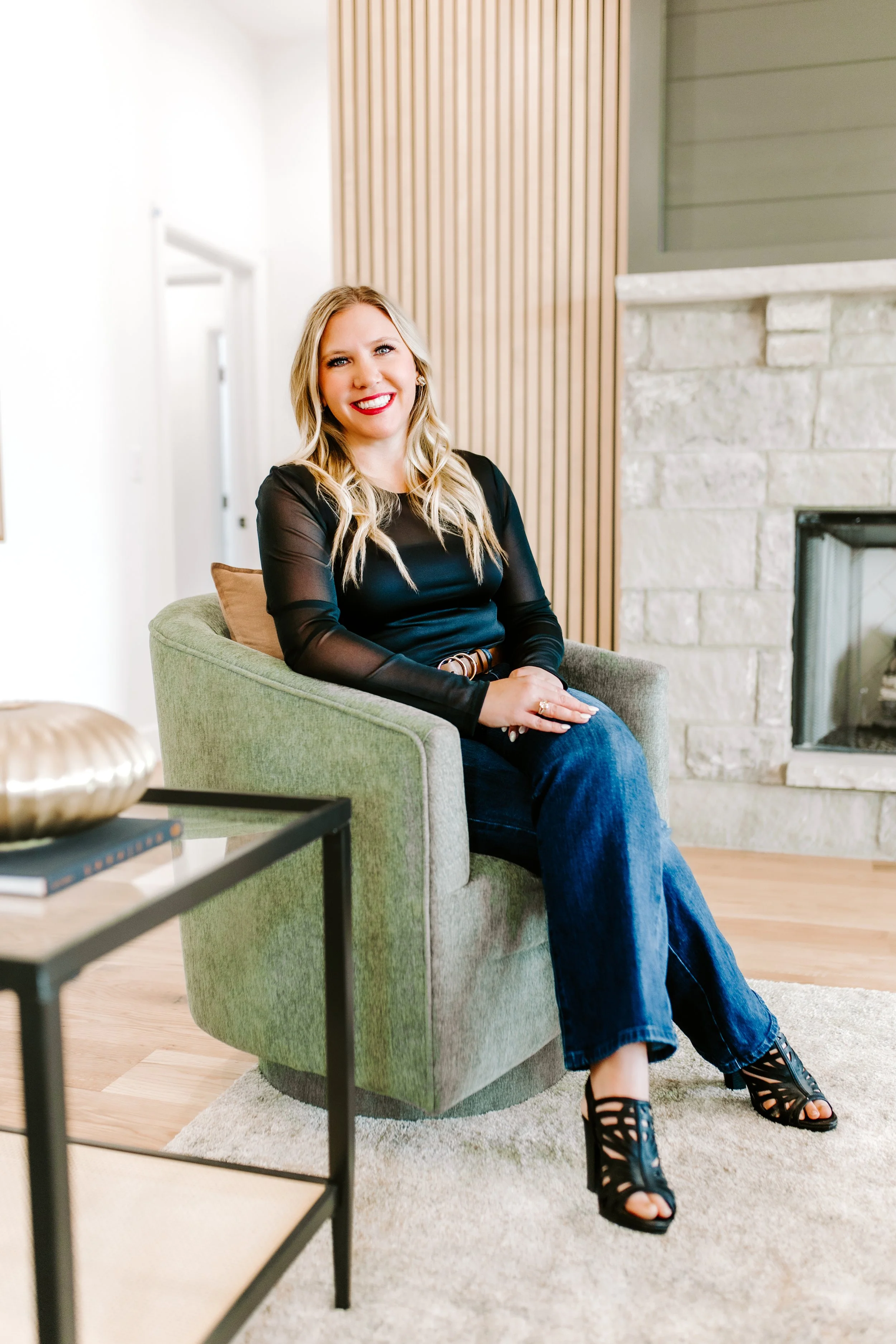 A woman with blonde hair and red lipstick sitting on a green armchair in a modern living room with a stone fireplace and wooden panel wall.