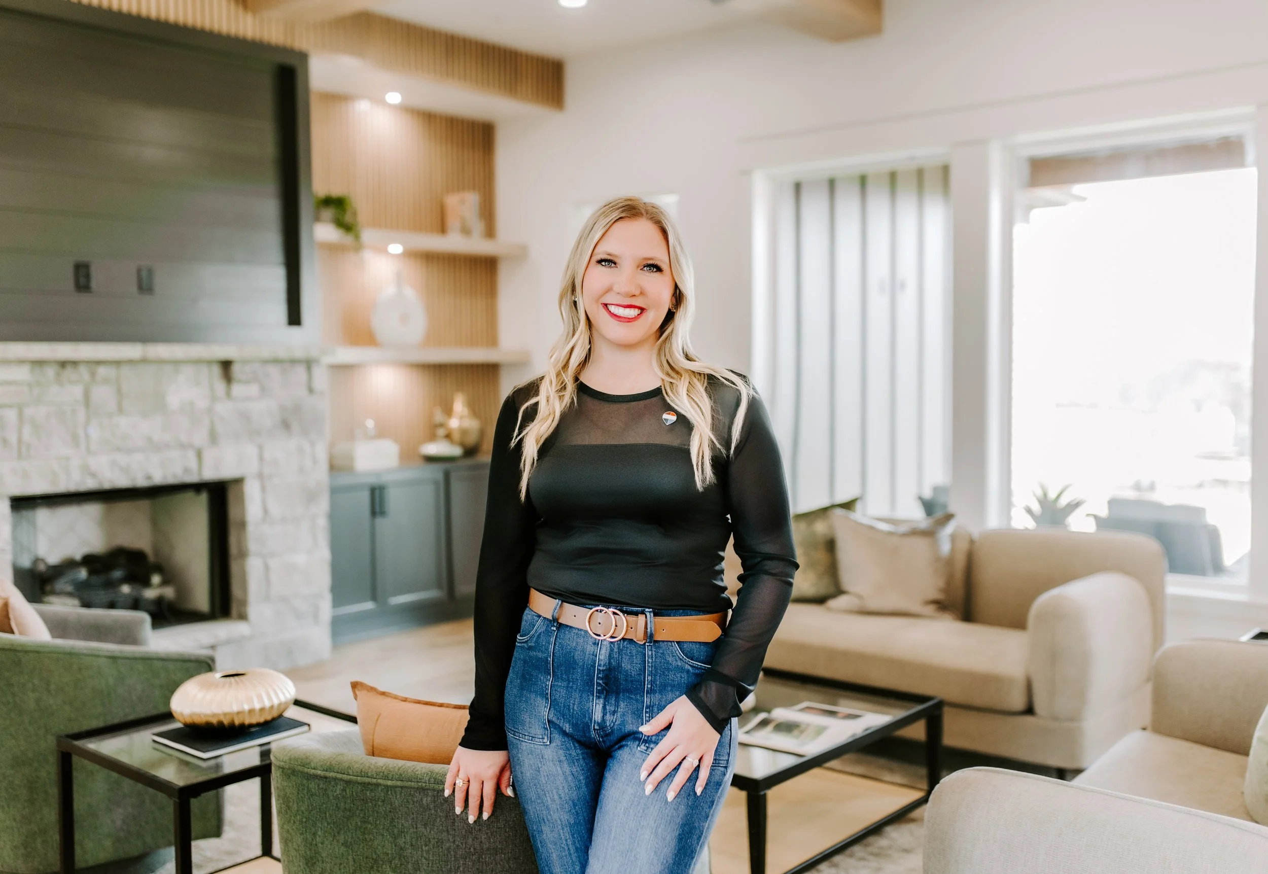 A woman wearing a black long-sleeve top with sheer detail and blue jeans, standing in a modern living room with a beige and green sofa, a black coffee table with decorative items, a fireplace, and large windows with vertical blinds.