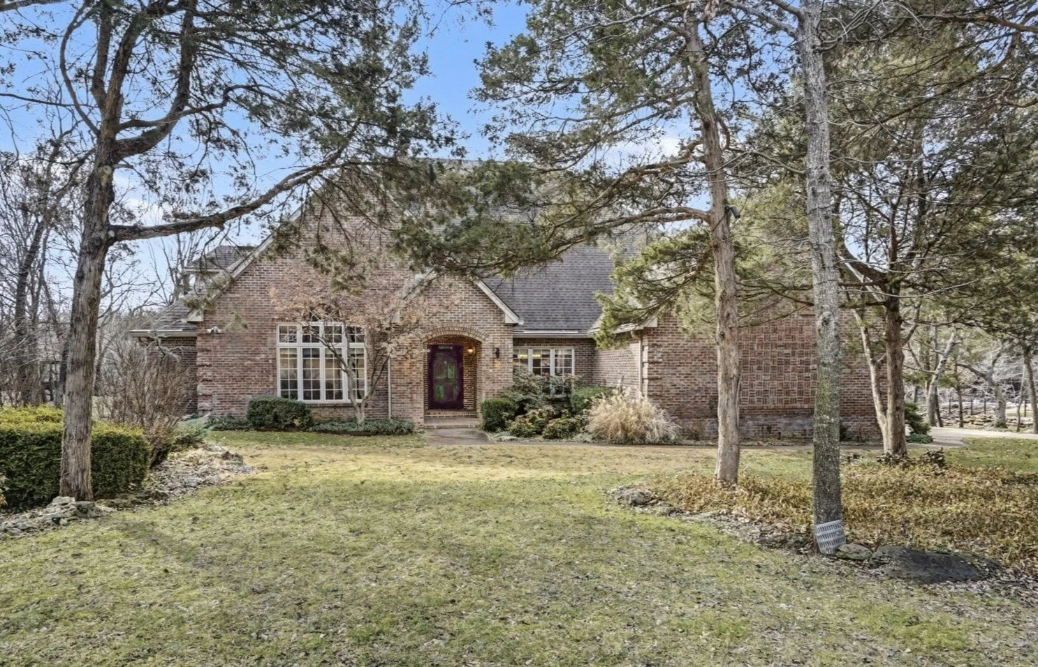 Brick house with large front yard, mature trees, and landscaped bushes, situated in a suburban neighborhood.