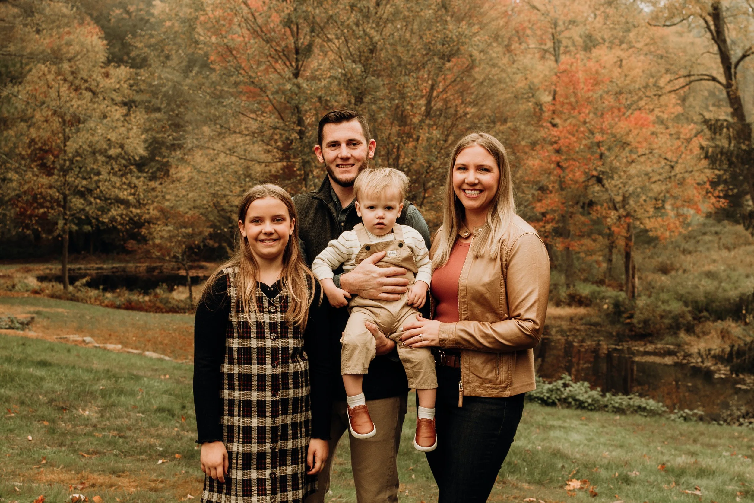 A family of four standing outdoors in front of autumn trees and a pond, smiling at the camera.
