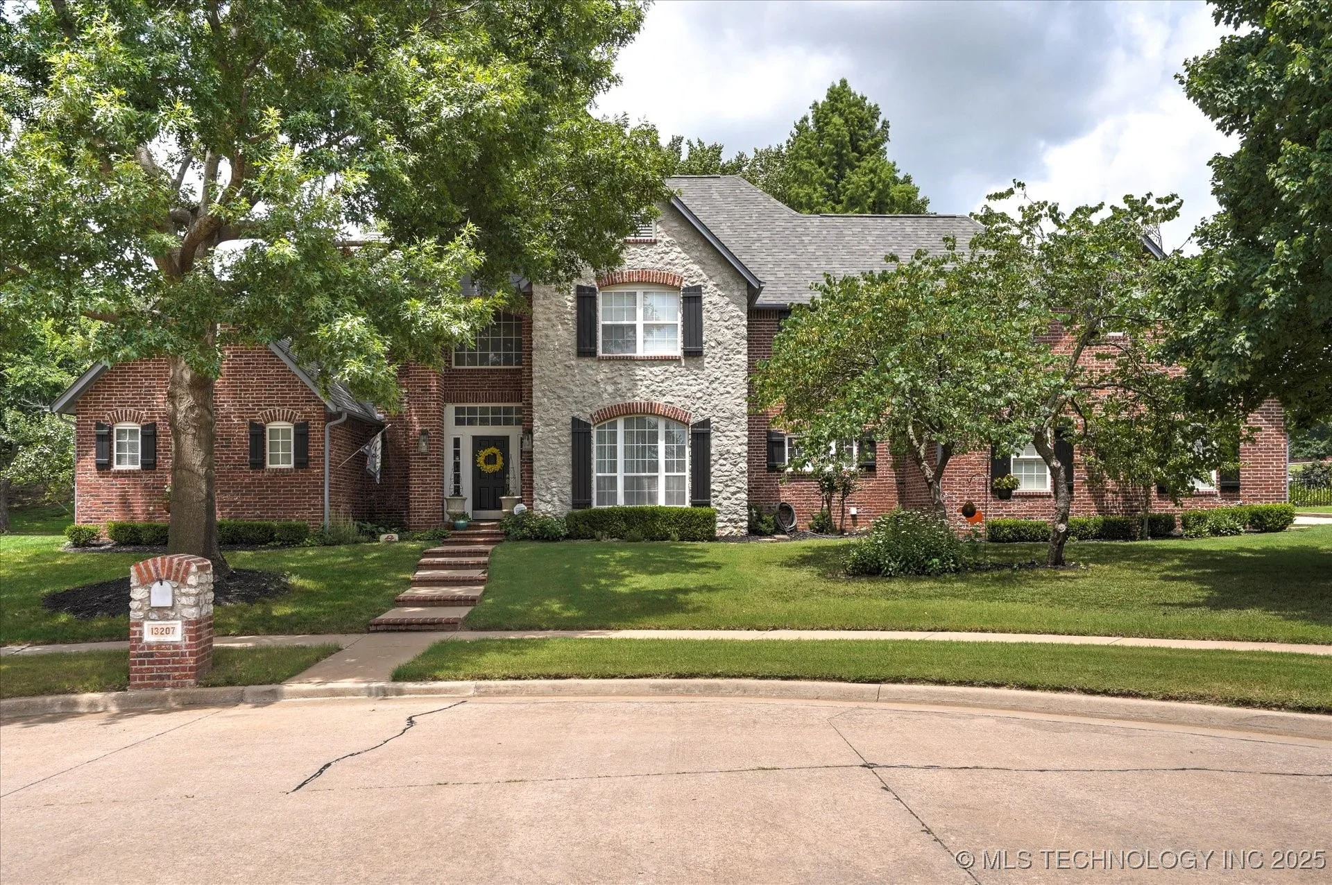 Two-story brick house with central white stone facade, black shutters, and a front lawn with trees, bushes, and a sidewalk.
