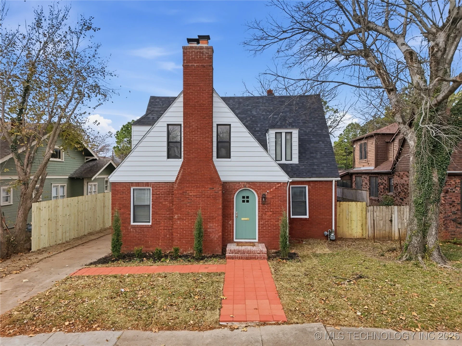 Front view of a house with brick and white siding, green door, small front yard, brick pathway, large trees, and neighboring houses surrounding it.