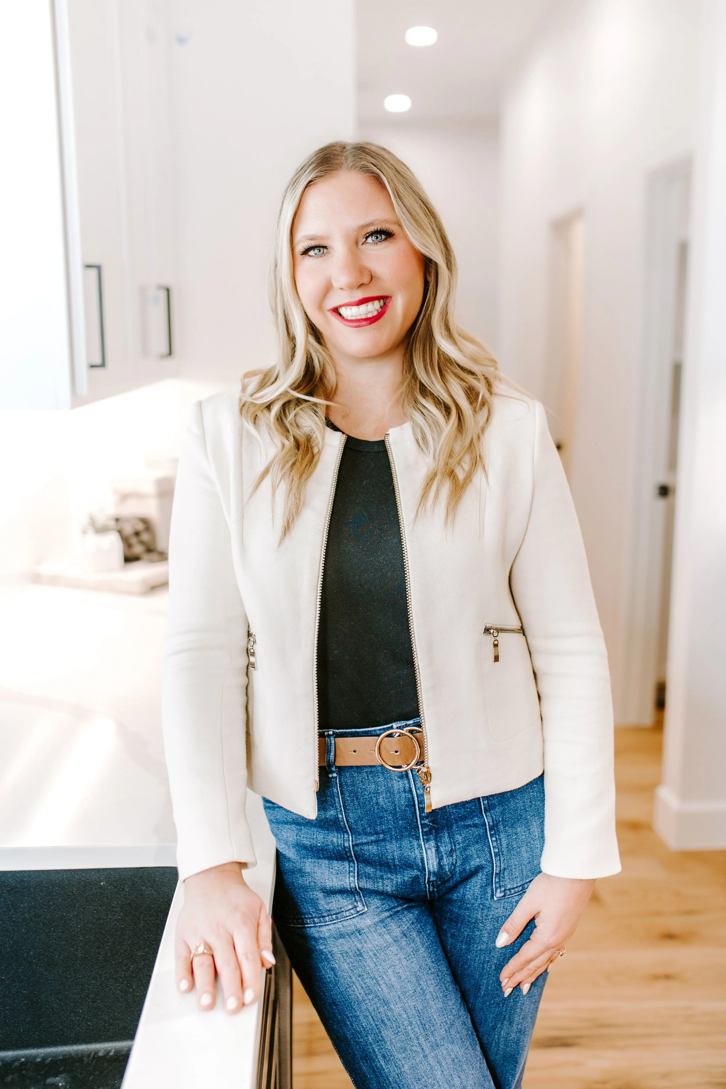 A smiling woman with blonde hair, wearing a cream-colored jacket, black top, and blue jeans, standing in a bright, modern home interior.