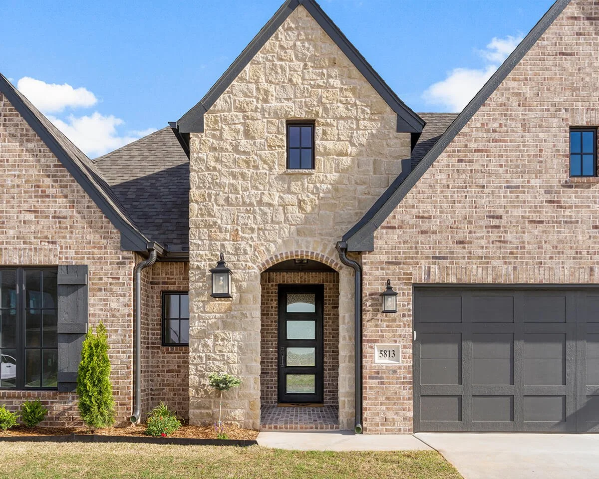 Front view of a brick house with a stone archway entrance, black front door, two windows, black shutters, a garage, outdoor lanterns, and small plants.