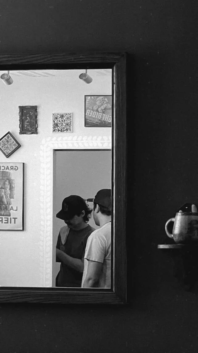 Two people wearing caps are reflected in a mirror, with one person showing the side profile and the other looking down at something in hand, inside an art or tattoo studio.